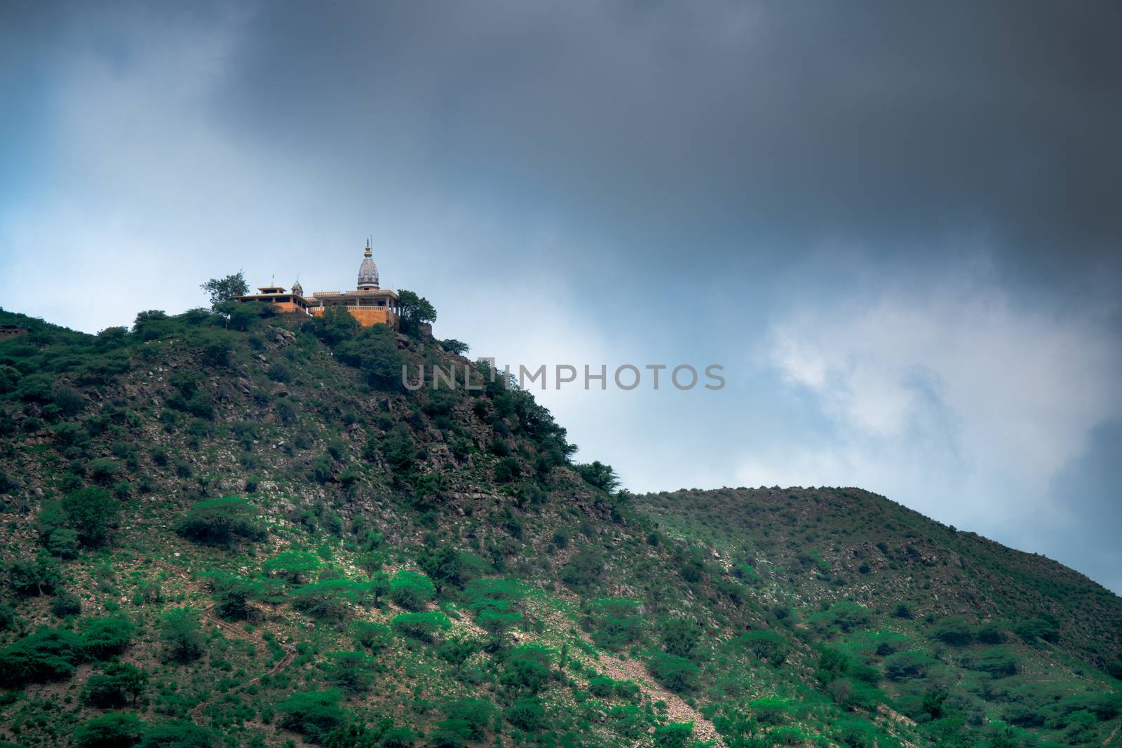 hindu temple on a hill covered in vegetation with the tower and spire clearly visible in the distance with a background of dark monsoon clouds shot in Rajasthan India by Shalinimathur