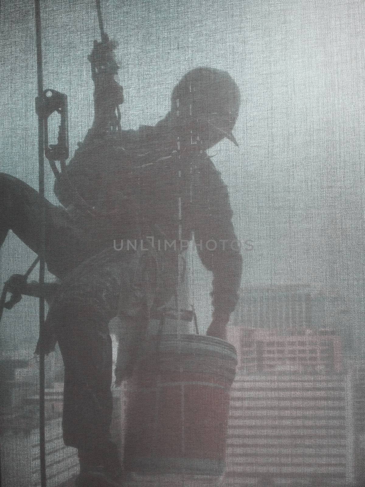 Silhouette images and shoot through transperent curtain effect from inside building which a man cleaning the window of high office building with his equipment such as wipe, sponge, bucket and high risk of danger