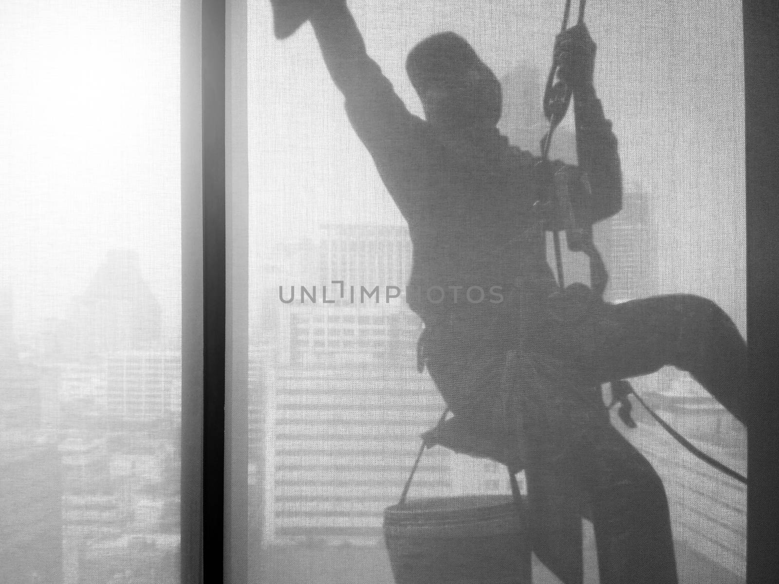 Silhouette images and shoot through transperent curtain effect from inside building which a man cleaning the window of high office building with his equipment such as wipe, sponge, bucket and high risk of danger