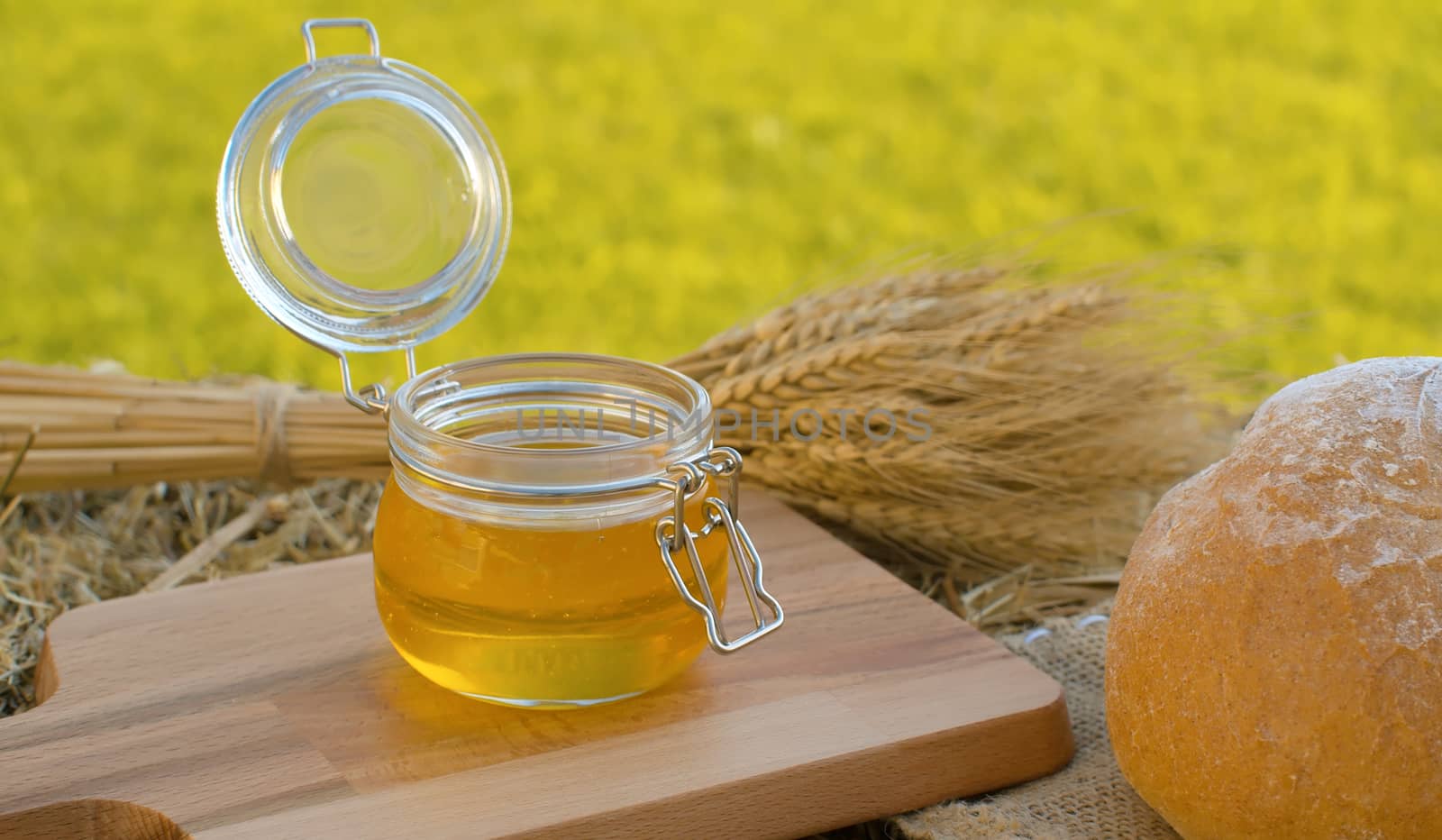 Close up honey in a glass jar. Traditional baked bread and sheaf of ripe rye ears on a fresh hay. Healthy food concept. Summer still life