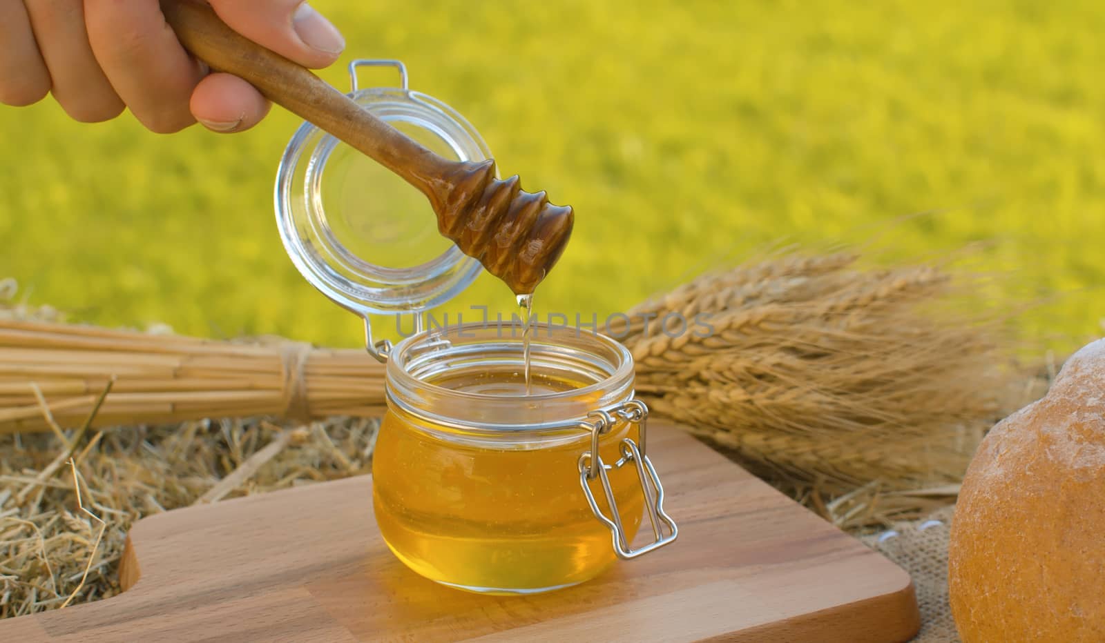 Close up hand taking honey with honey spoon. Traditional baked bread and sheaf of ripe rye ears on a fresh hay. Healthy food concept. Summer still life