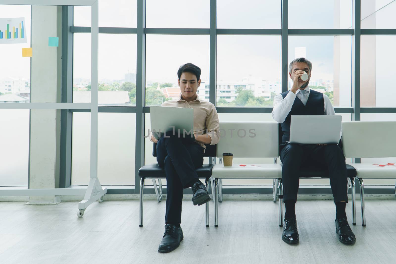 group of businessmen using laptops and drinks coffee before a business meeting. Asian young men and Adult male Caucasian people talking while awaiting an interview. social distancing is new normal