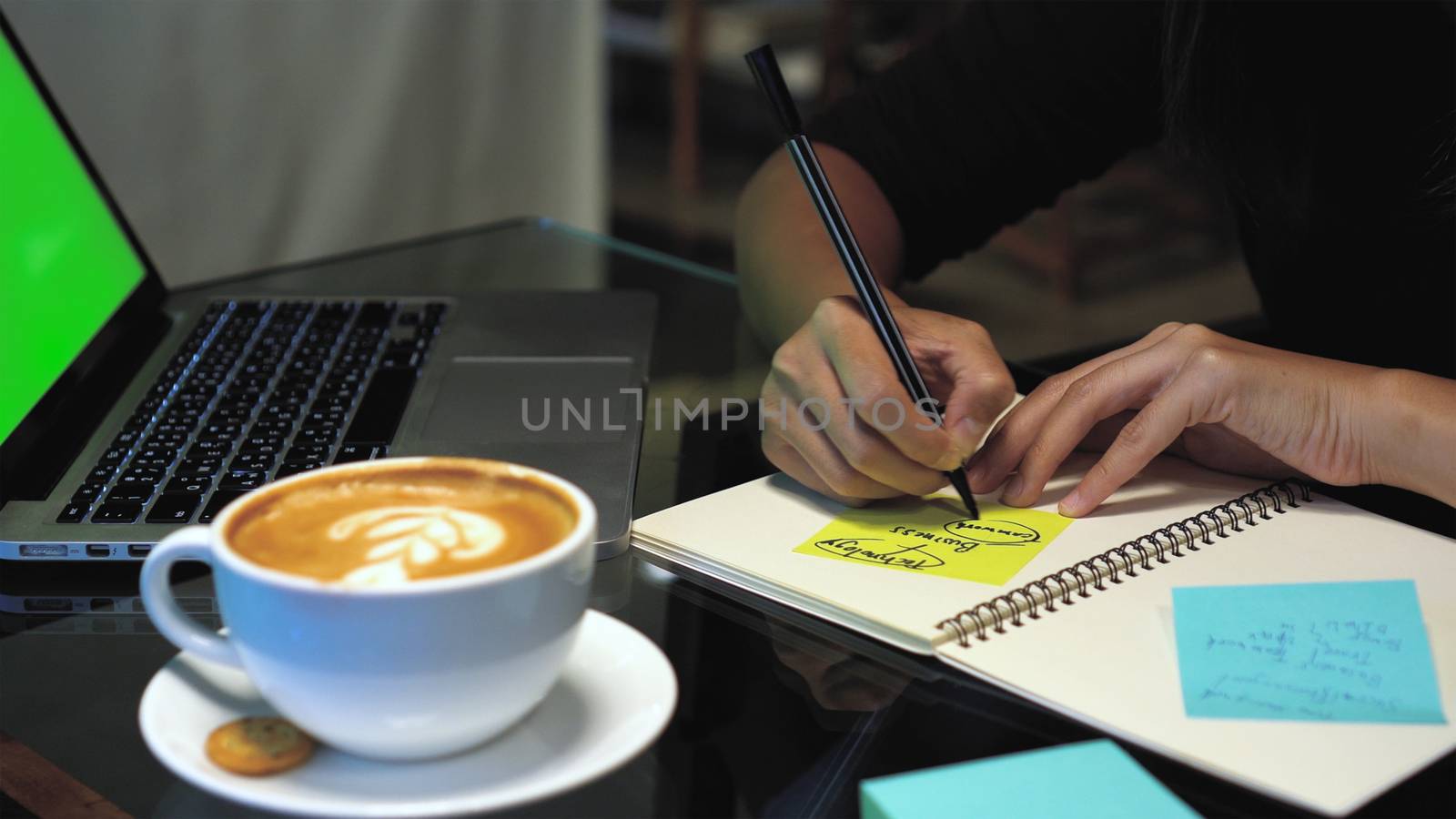 Closeup hands of Asian businesswoman working and writing on paper Notes over the notebook with green screen laptop and coffee cup in coffee cafe, Lifestyle and business concept