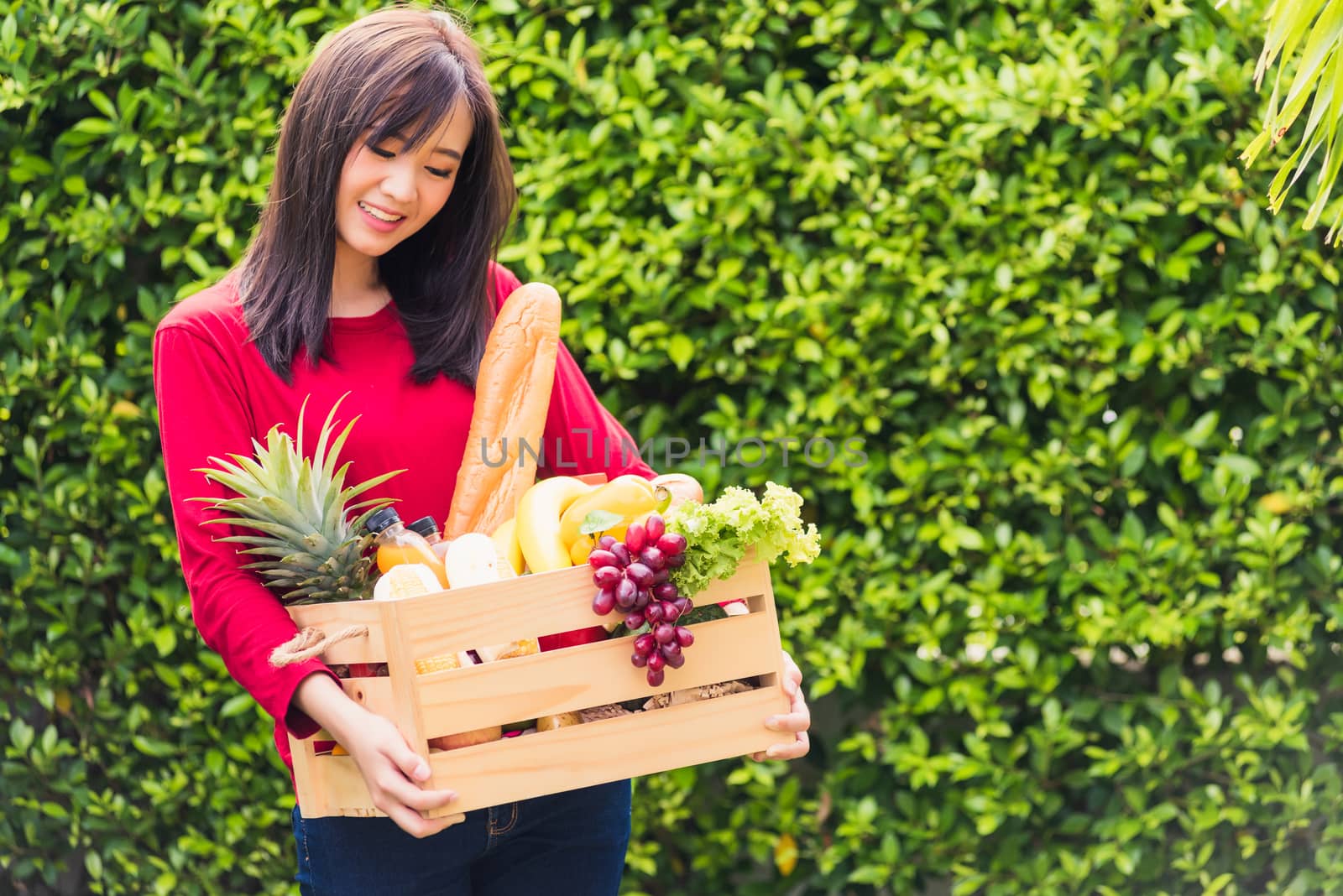 Portrait of Asian beautiful young woman farmer standing she smile and holding full fresh food raw vegetables fruit in a wood box in her hands on green leaves background