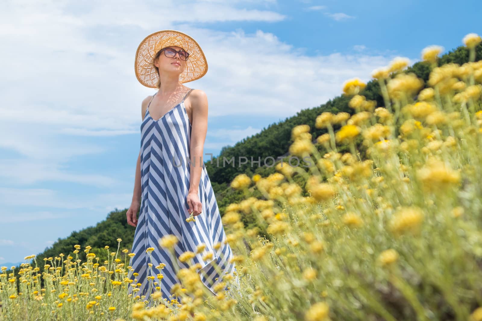 Young woman wearing striped summer dress and straw hat standing in super bloom of wildflowers, relaxing while enjoing beautiful nature of of Adriatic sea coastal nature of Croatia.