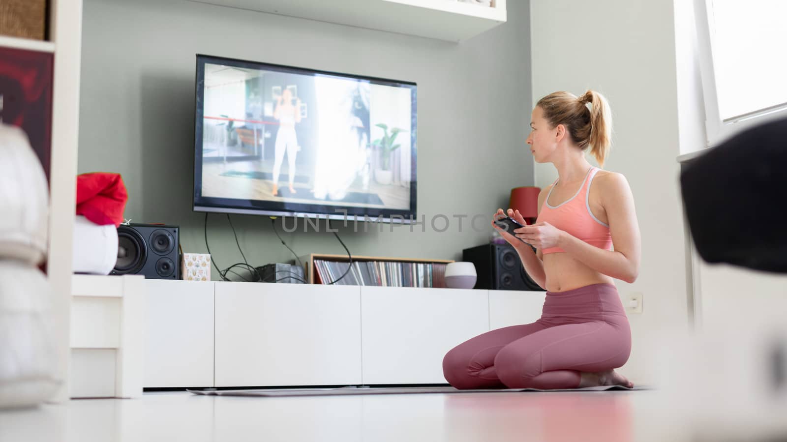 Attractive sporty woman working out at home, doing pilates exercise in front of television in small studio appartment. Social distancing. Stay healthy and stay at home during corona virus pandemic.