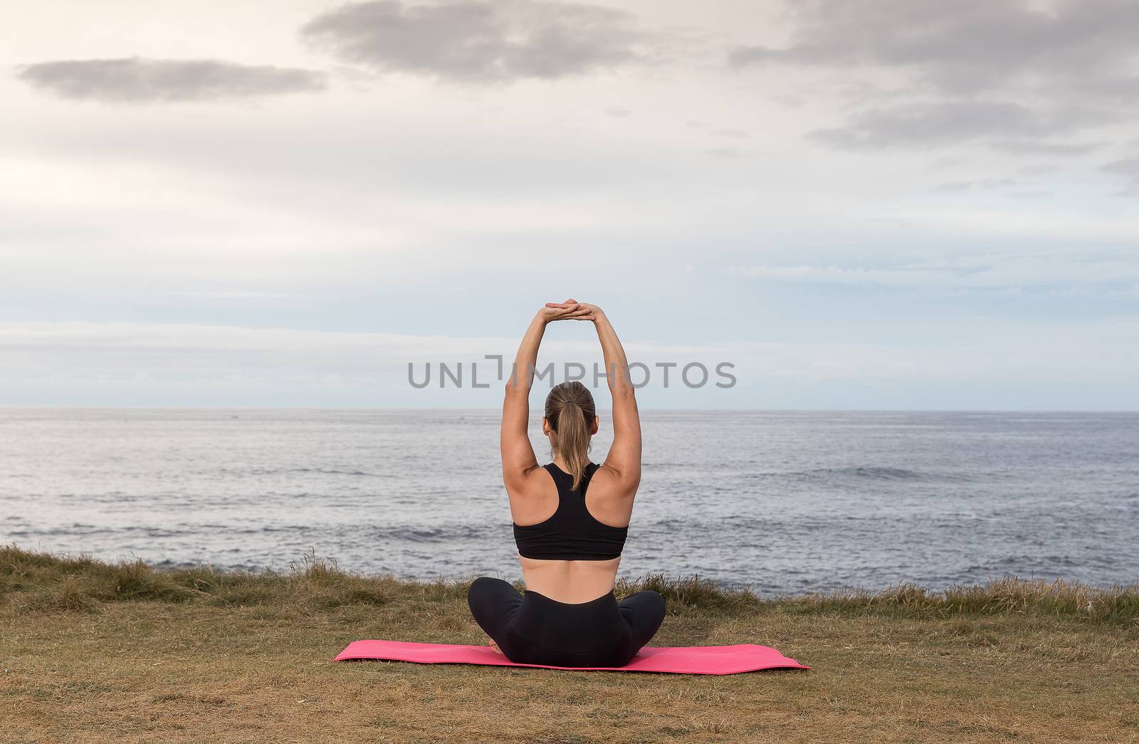 Woman in black sportswear exercising outdoors on a pink mat with the sea in the background.