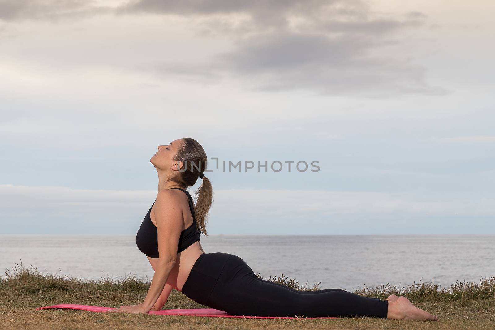 Woman in black sportswear doing pilates outdoor on a pink mat with the sea in the background.