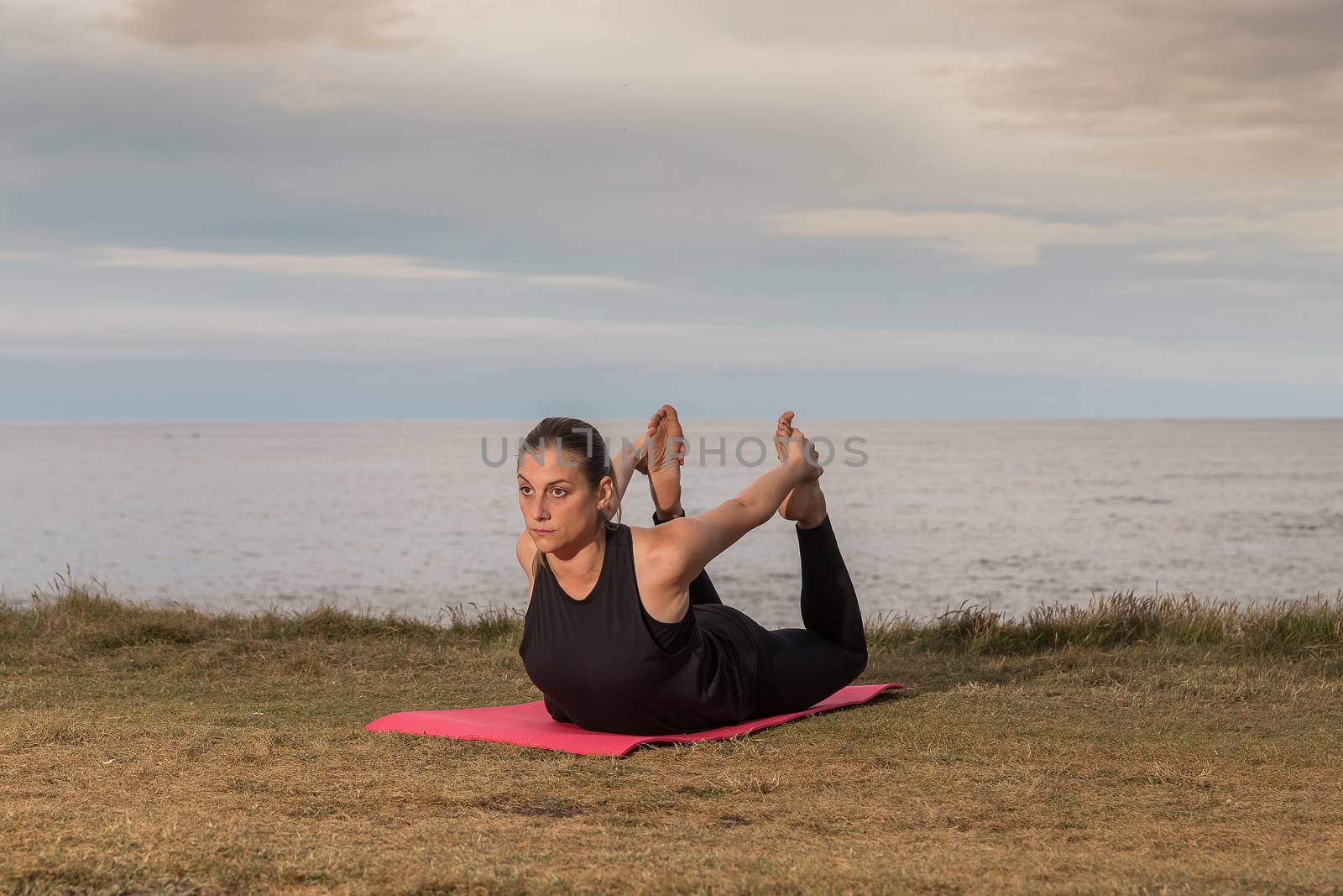 Woman in black sportswear exercising outdoors on a pink mat with the sea in the background.