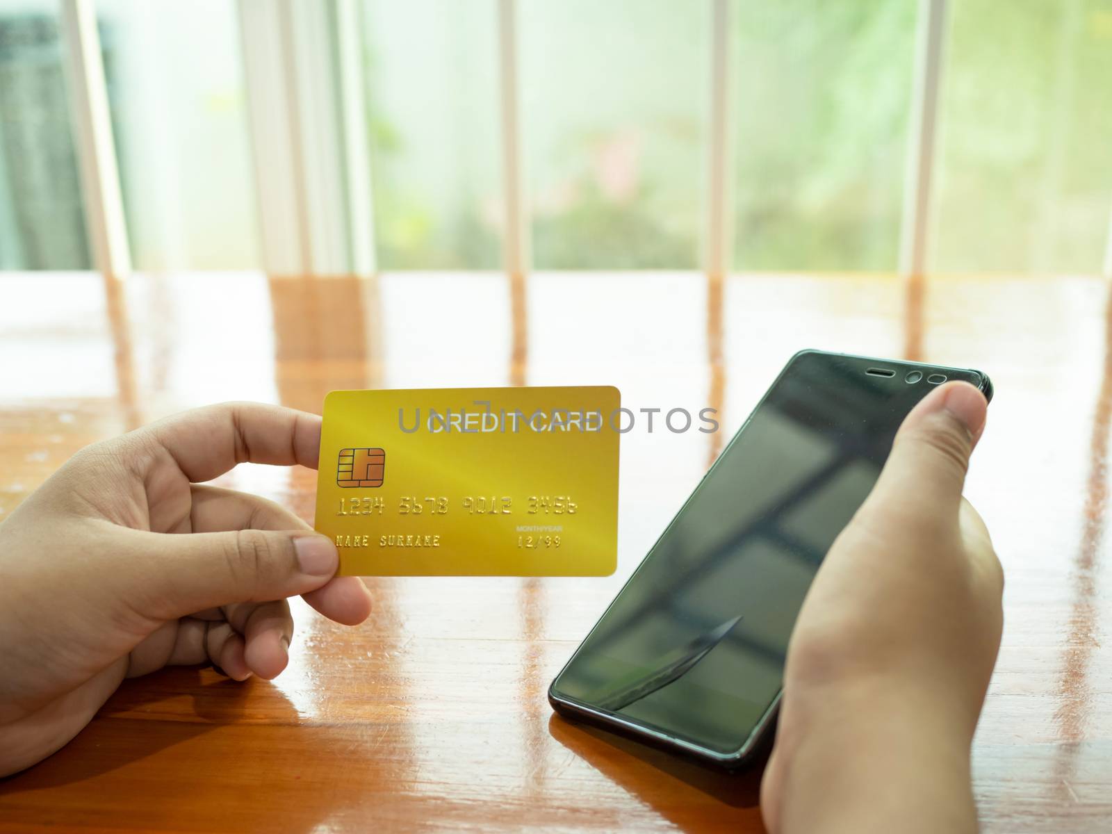 Online shopping concept with human hand holding smart phone and credit card on wood table and window background.