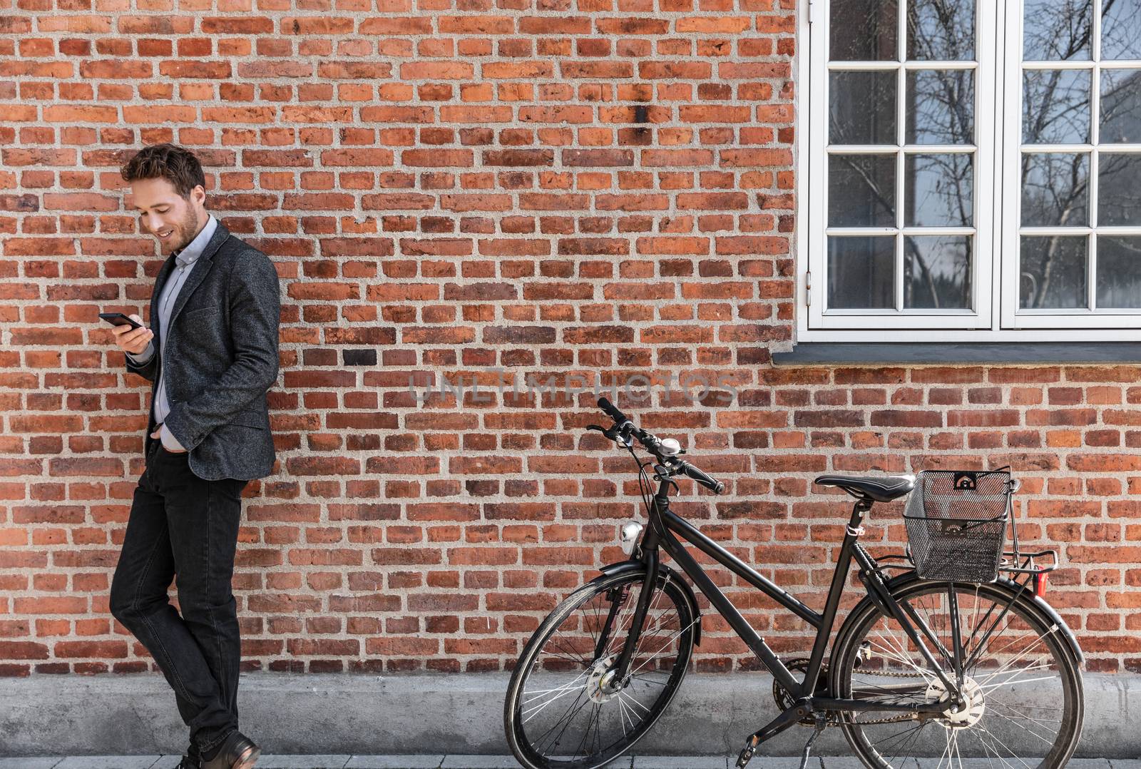 Young businessman going to work on bike commute using his mobile cellphone against city brick wall background. Happy business man bicycle commuter arriving at office using phone texting.