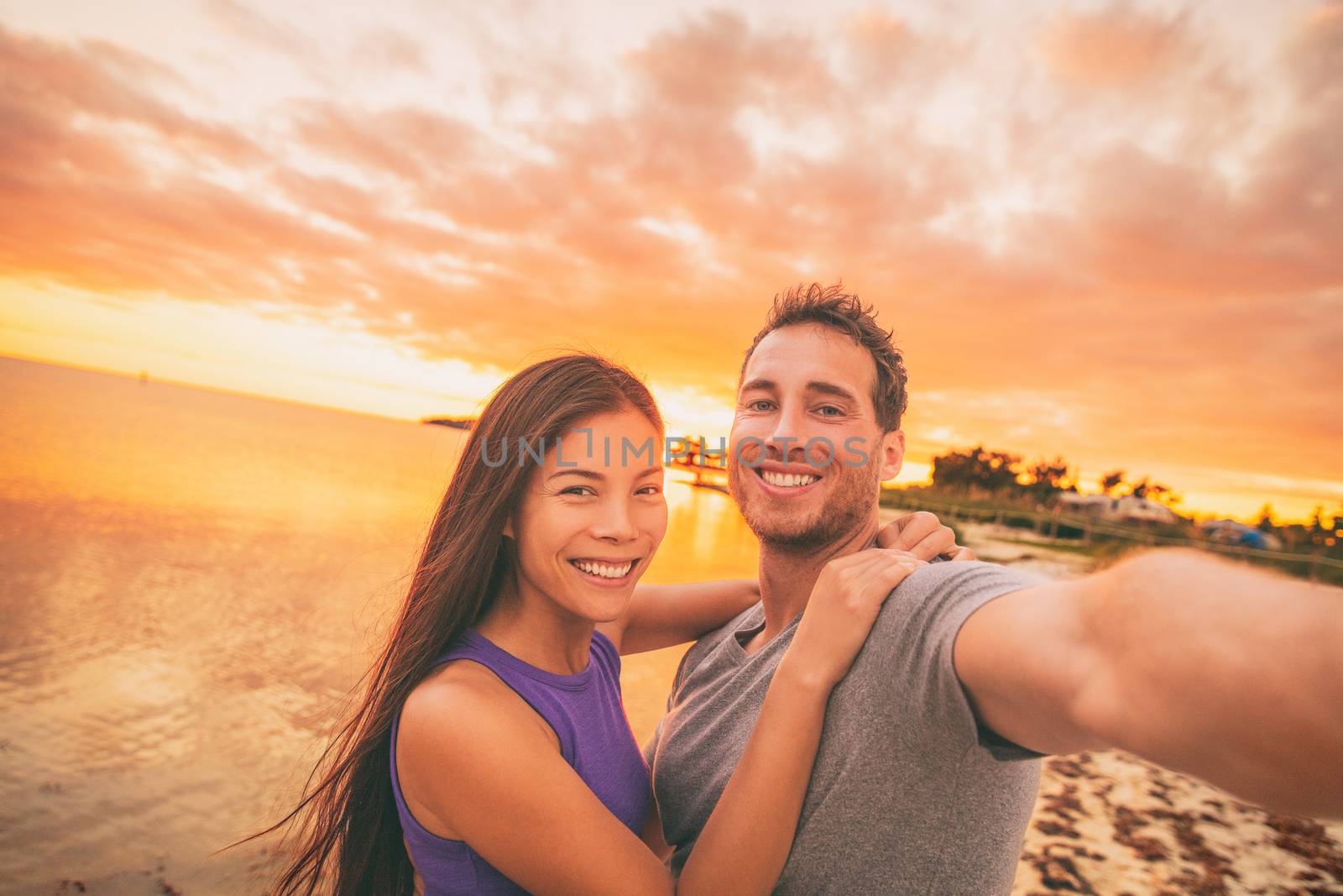 Happy selfie couple tourists on USA travel taking photo at sunset on Florida beach. Smiling Asian woman and Caucasian man, interracial relationship.