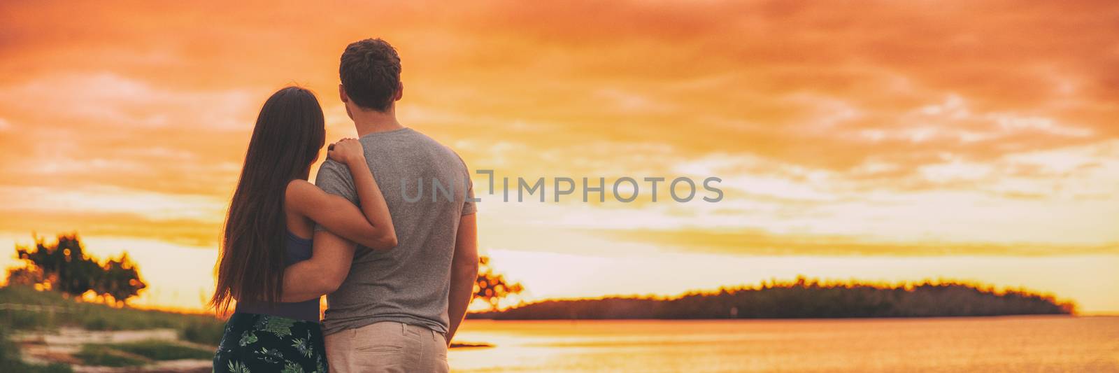 Couple watching sunset on summer adventure travel at beach panoramic banner - glow sky background at dusk.