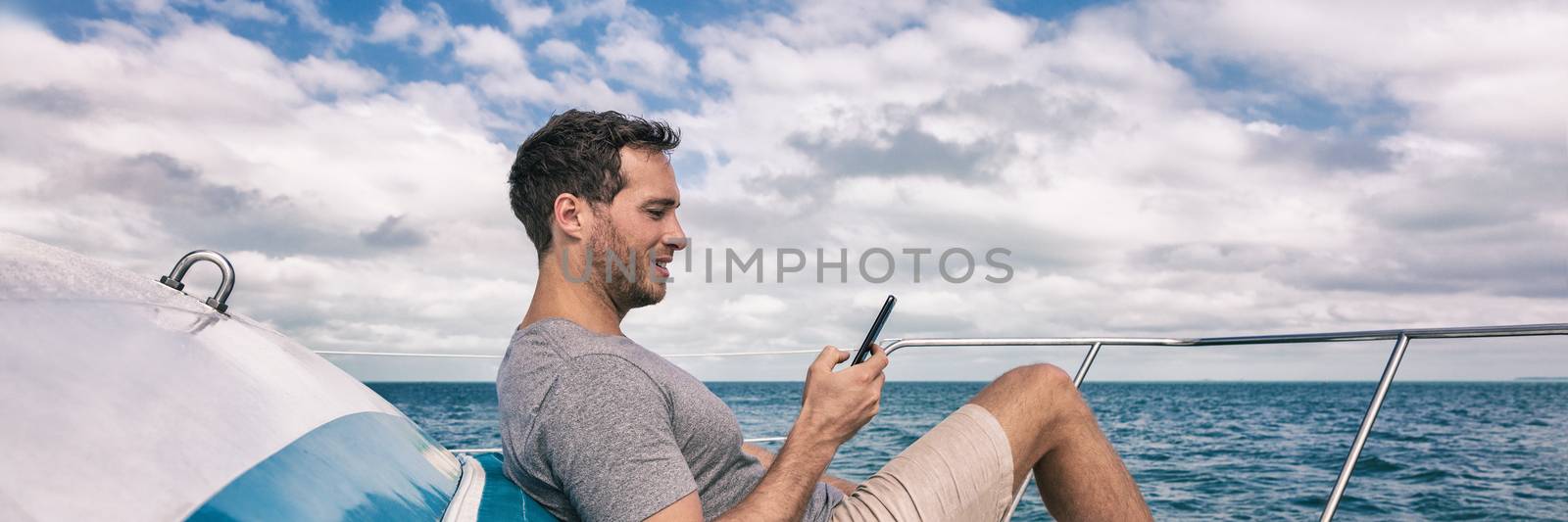 Yacht luxury lifestyle young man using cellphone banner panorama. Person relaxing on deck texting sms message on mobile phone under the sun summer holidays by Maridav