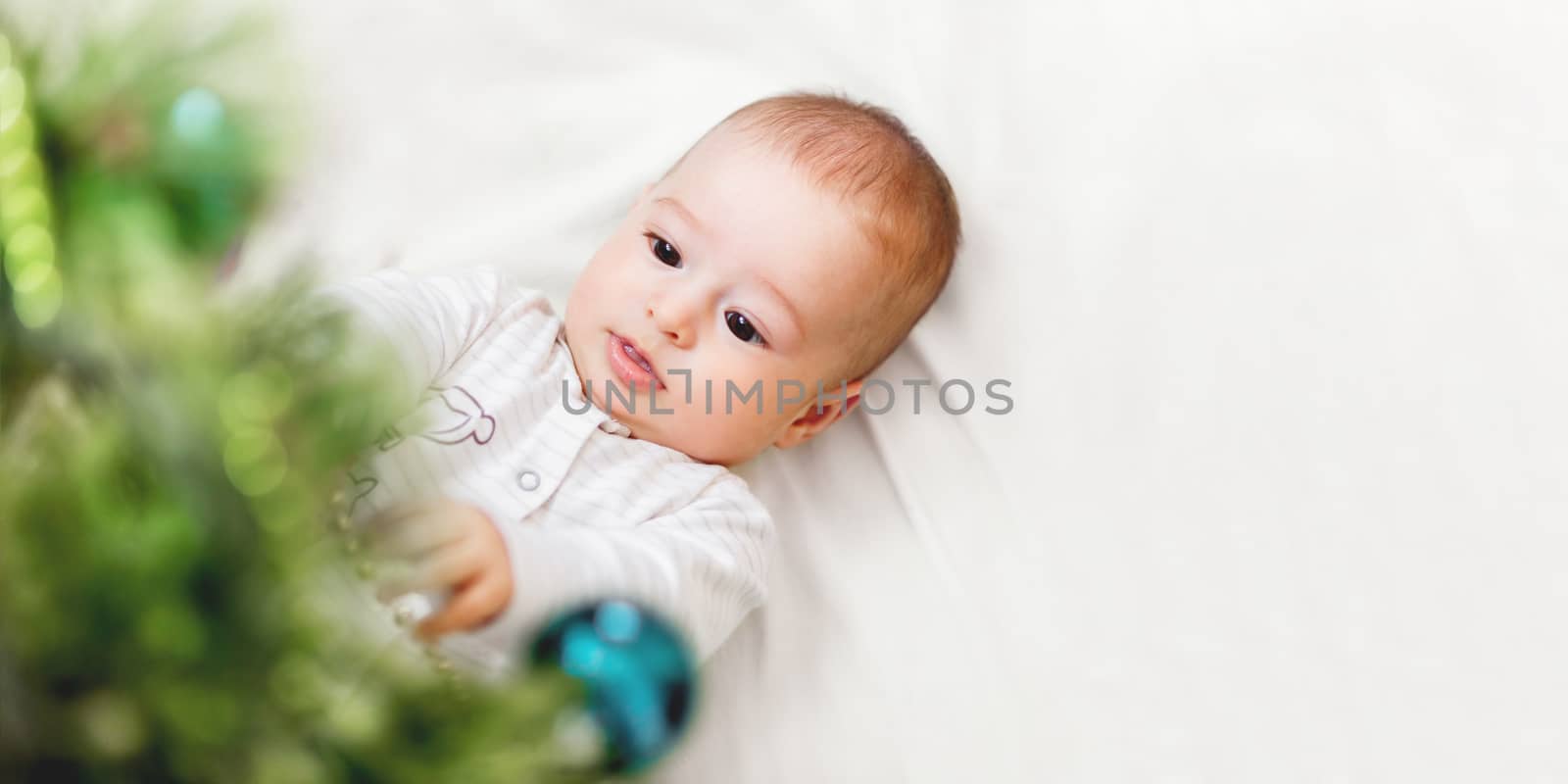 Baby boy or girl lying under Christmas tree. Little child plays with fir tree decorated for New Year celebration. Cute kid portrait. Winter holiday spirit. Banner with copy space.