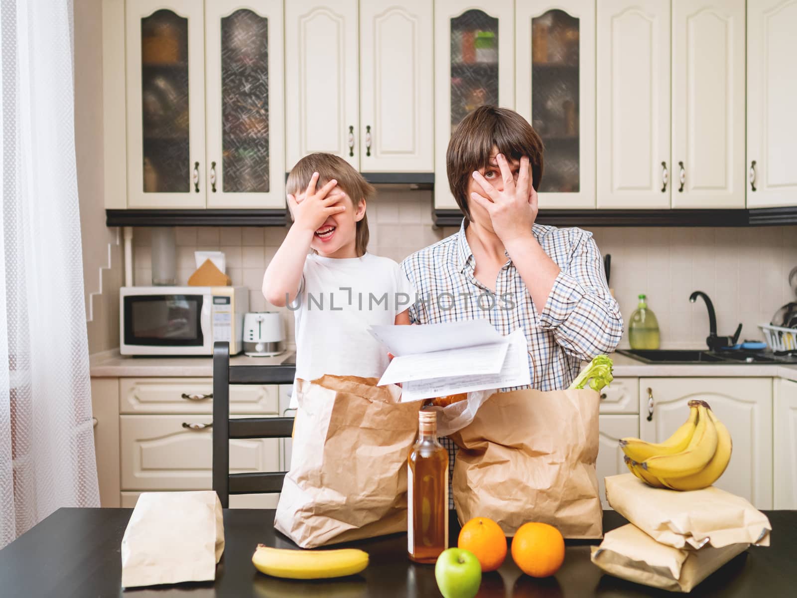 Family sorts out purchases in the kitchen. Father and son shows expensiveness of products in shopping bags. Grocery delivery in conditions of quarantine because of coronavirus COVID19.