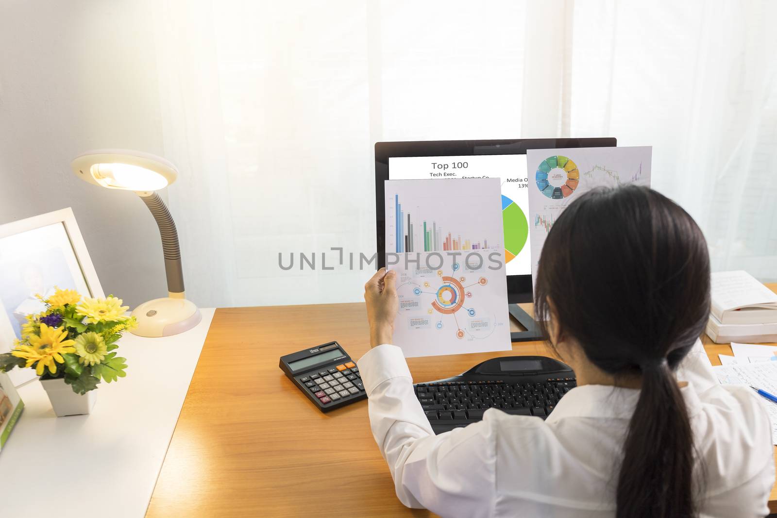 Rear view of business woman working in office with computer holding chart report paper and looking. business people working at home with paper and PC screen. Business and finance , work at home concept.