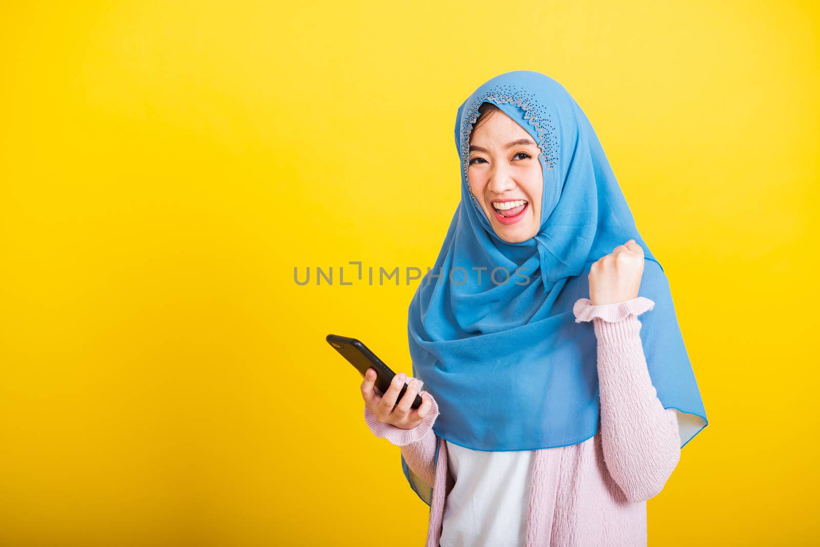 Asian Muslim Arab, Portrait of happy beautiful young woman Islam religious wear veil hijab funny smile she raise hand glad celebrating winning with smart mobile phone isolated on yellow background