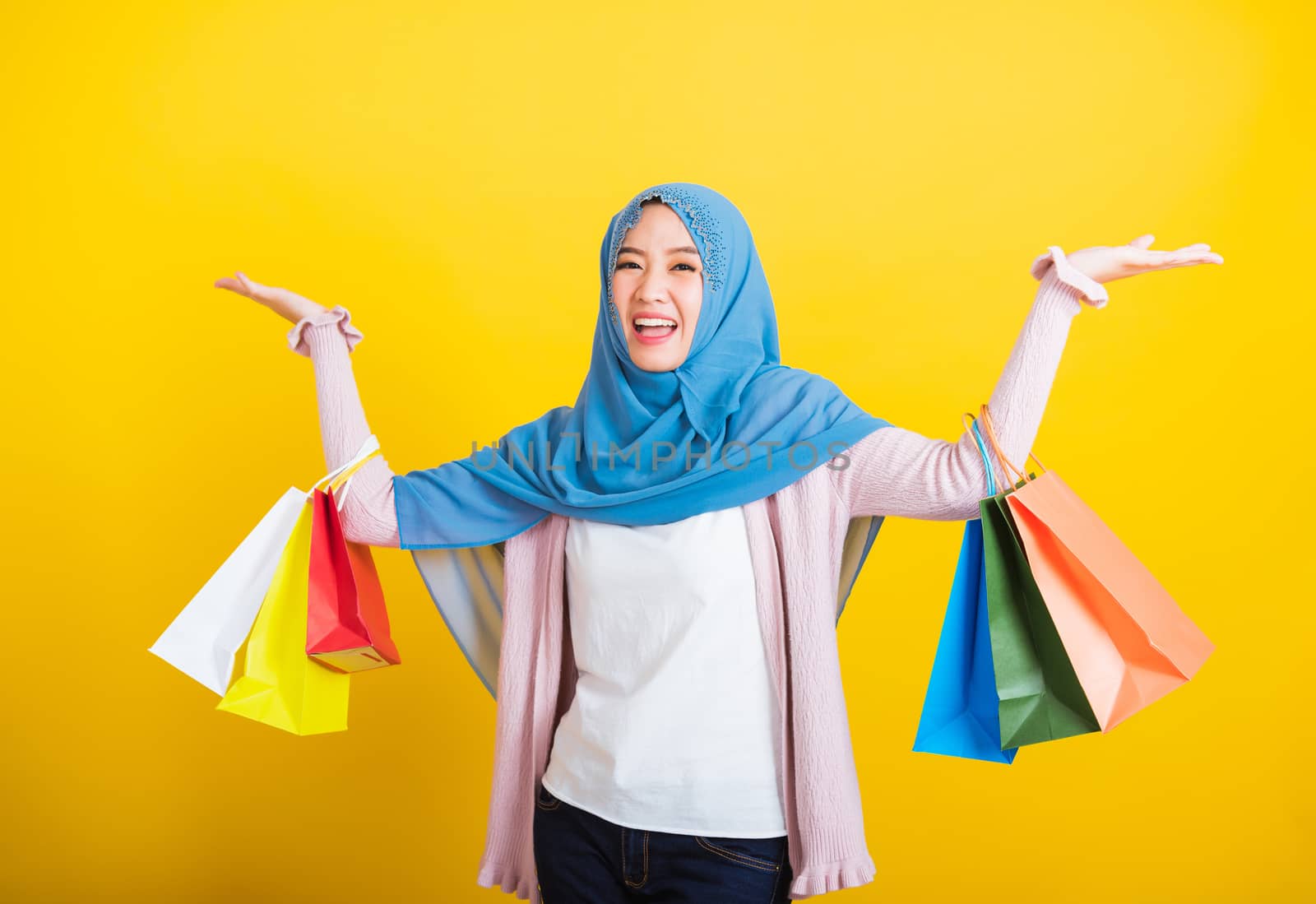 Asian Muslim Arab, Portrait of happy beautiful young woman Islam religious wear veil hijab funny smile she holding colorful shopping bags so glad shopping bags hand raise in studio isolated on yellow