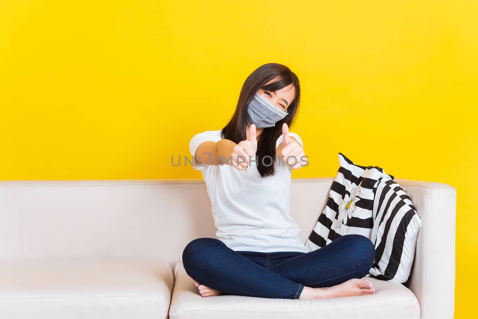 Portrait Asian of beautiful young woman sitting on sofa wearing medical face mask protective show finger thumb up for good sign during Coronavirus studio shot isolated on yellow background