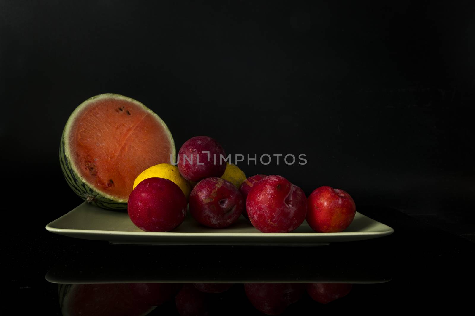 still life with a green bowl on a reflective background with lemons, plums and half a water meleon