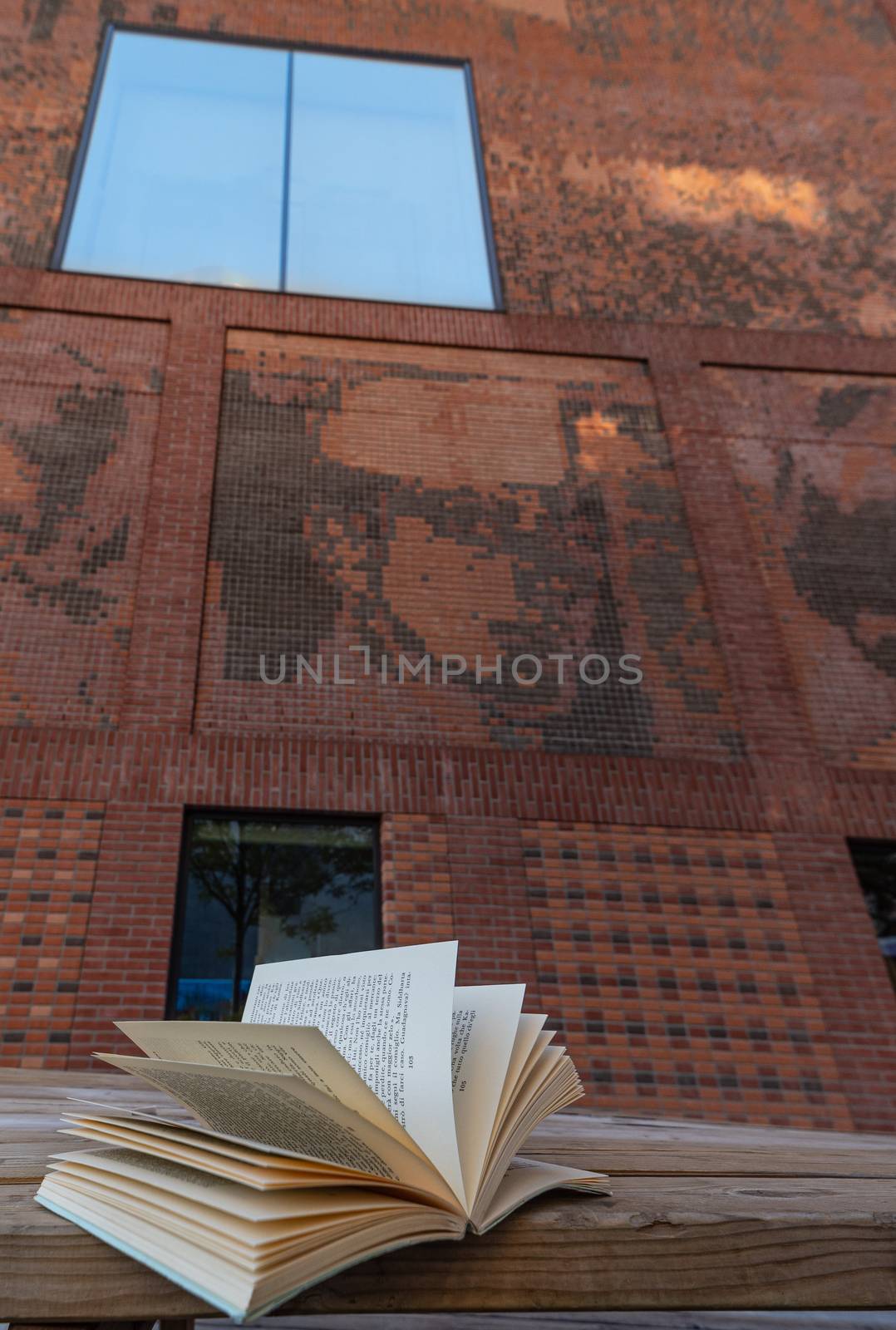 An open book resting on a bench in front of a red brick building with a stained glass window reflecting the blue sky