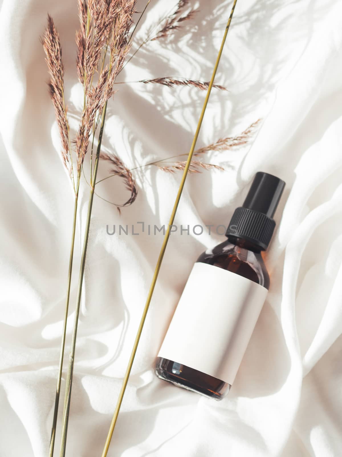 Concept of apothecary cosmetic. Herbs and brown glass bottle of essential oil in still life composition on crumpled white fabric. Sunlight and shadow. Natural cosmetic remedy with white tag.