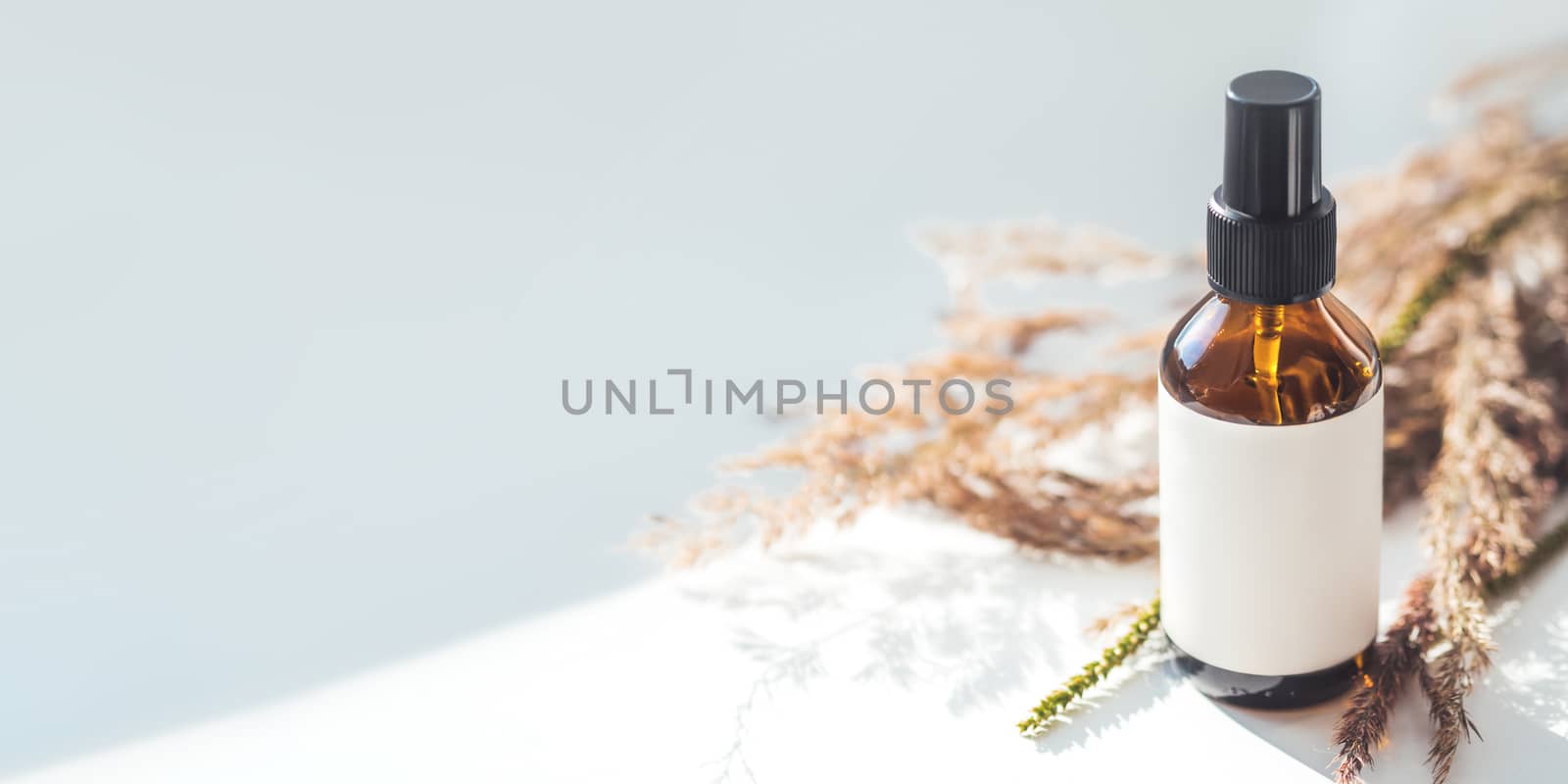 Concept of apothecary cosmetic. Herbs and brown glass bottle of essential oil in still life composition on light blue and white background. Sunlight and shadow. Natural cosmetic remedy with white tag.