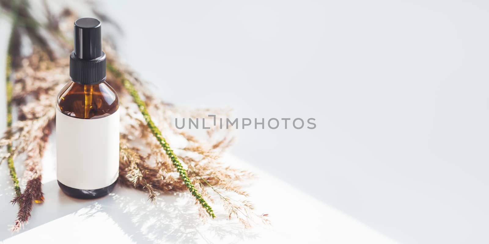 Concept of apothecary cosmetic. Herbs and brown glass bottle of essential oil in still life composition on light blue and white background. Sunlight and shadow. Natural cosmetic remedy with white tag.