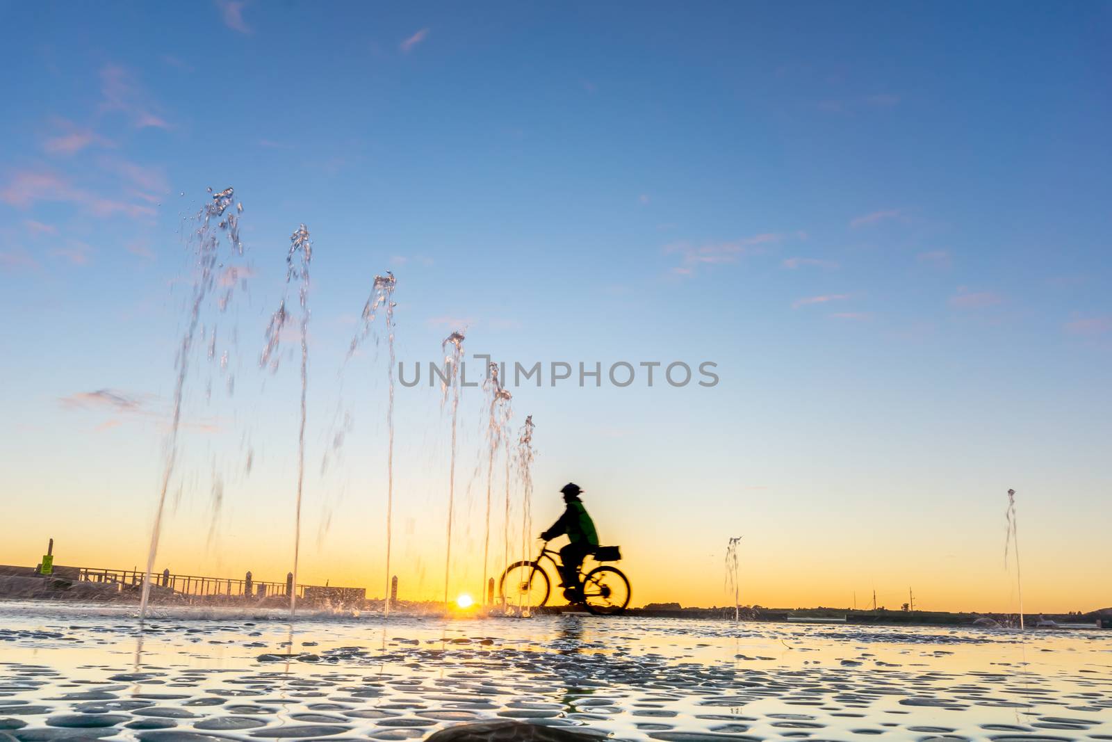 Water feature with spraying water on Tauranga Strand waterfront at sunrise with clear blue sky and blurred silhouette of cyclist passing in distance, New Zealand