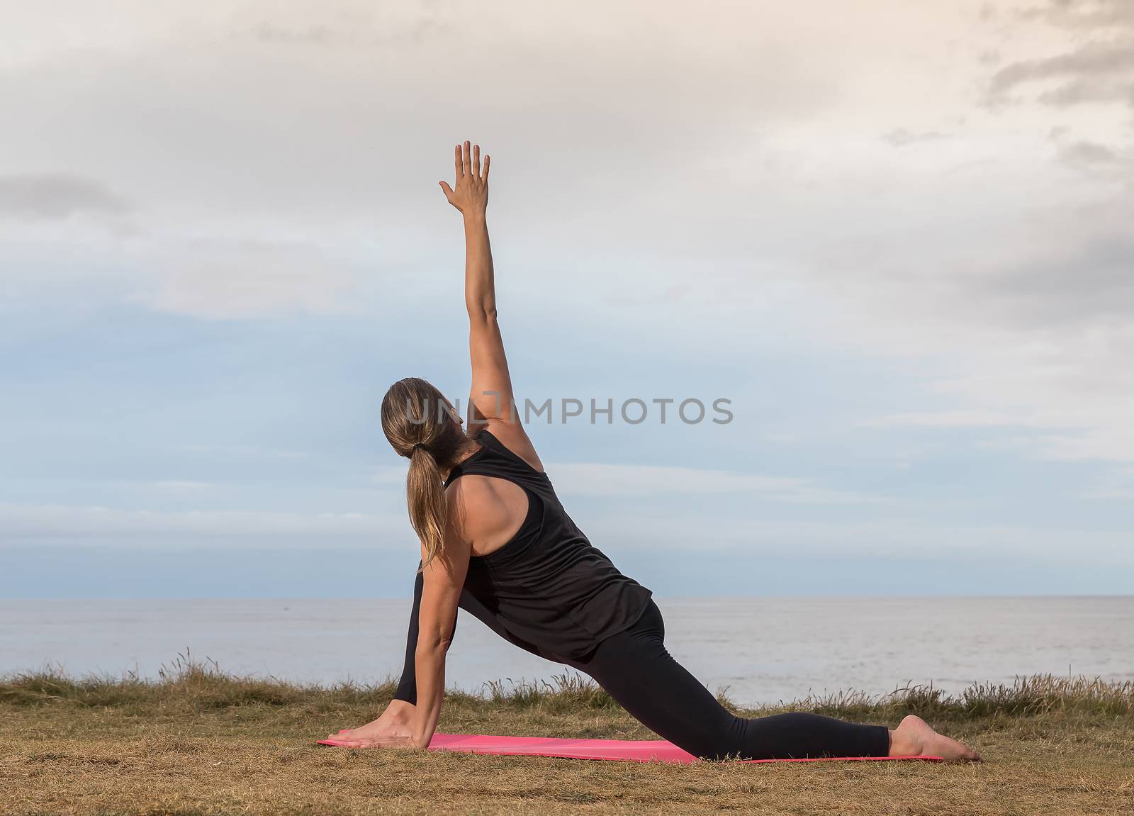 Woman in sportswear stretching outdoors on a pink mat with the sea in the background.