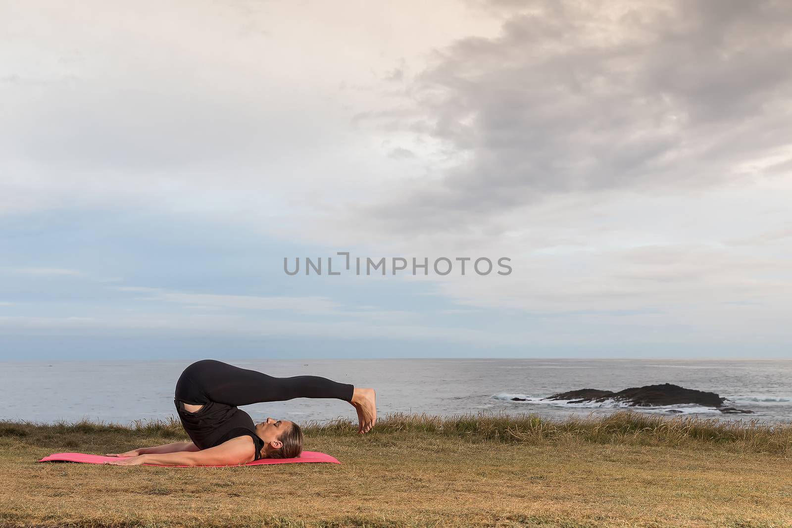 Woman in sportswear doing pilates outdoors on a pink mat with the sea in the background.