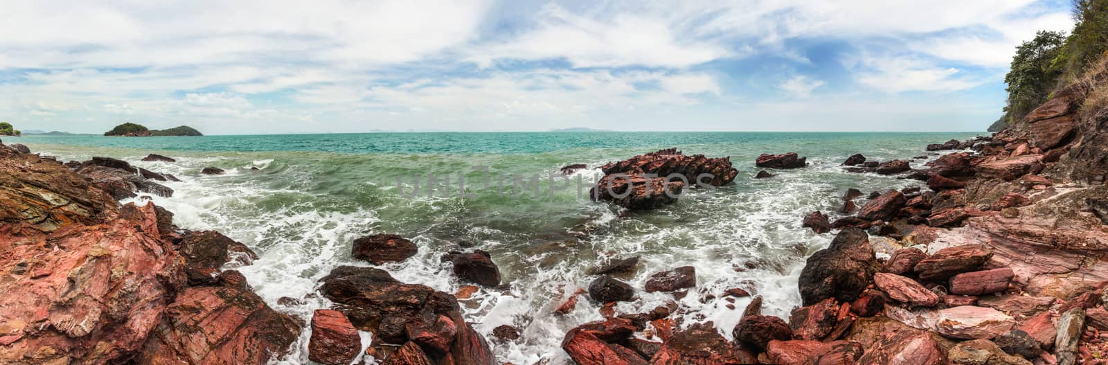 High resolution wide panorama of sea waves crushing on red rocks with blue sky with clouds in background. Koh Lanta, Thailand