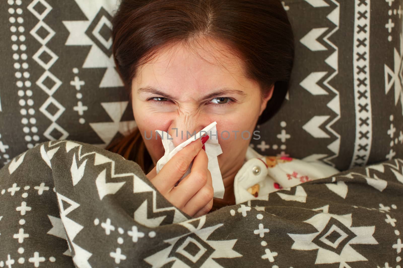 Sick young woman in pyjama, laying in bed, covered under duvet, holding tissue handkerchief in her hand to blow runny nose.