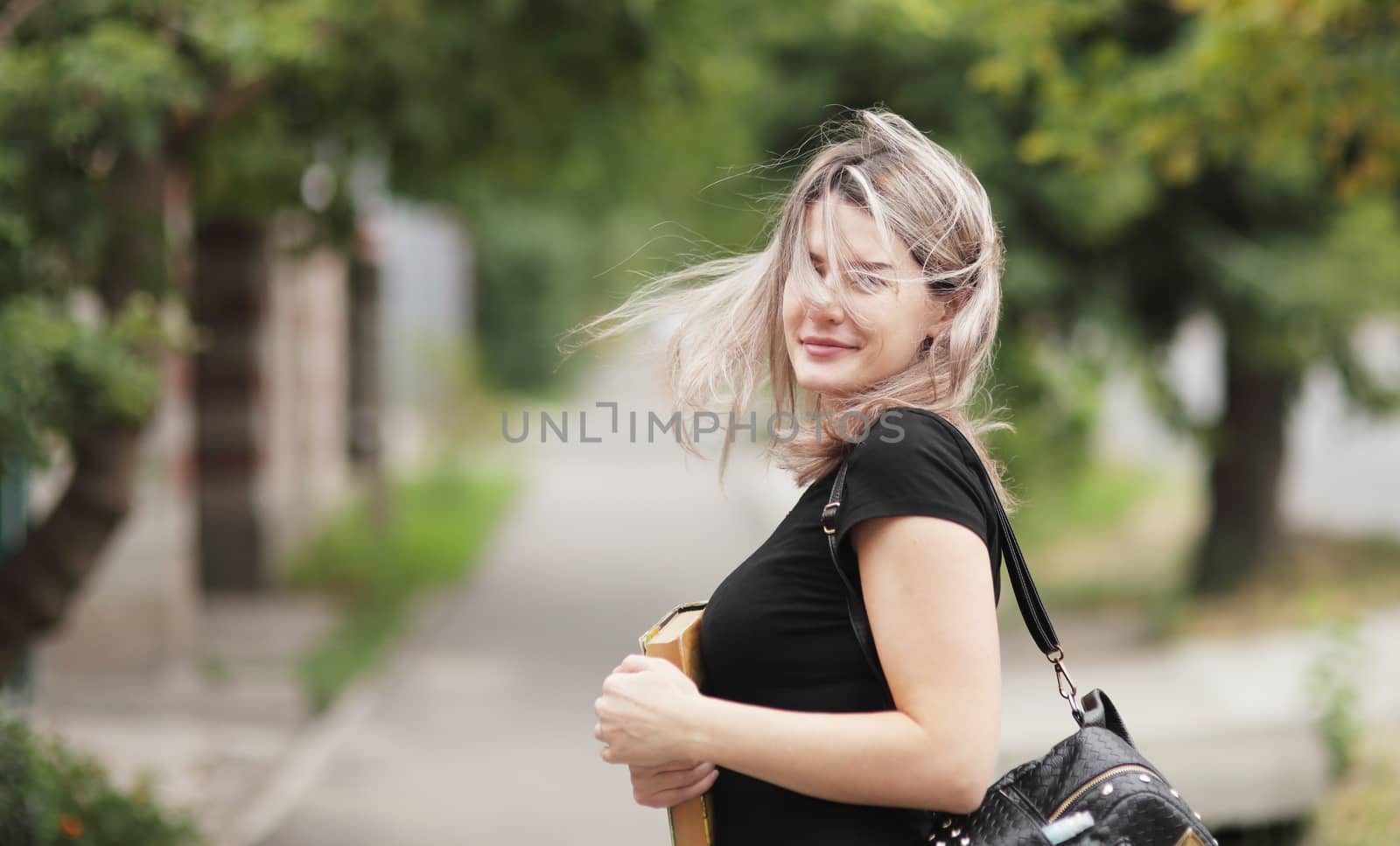 Fashion portrait of young stylish woman with fluttering hair walking down the street, in trendy outfit, traveling with a backpack. Holds a book in his hand