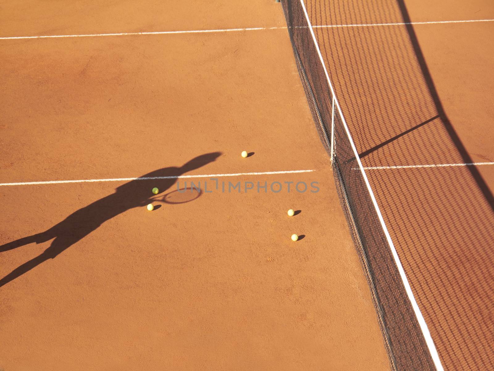 Shadow of tennis player at net with scatttered tennis balls on clay court