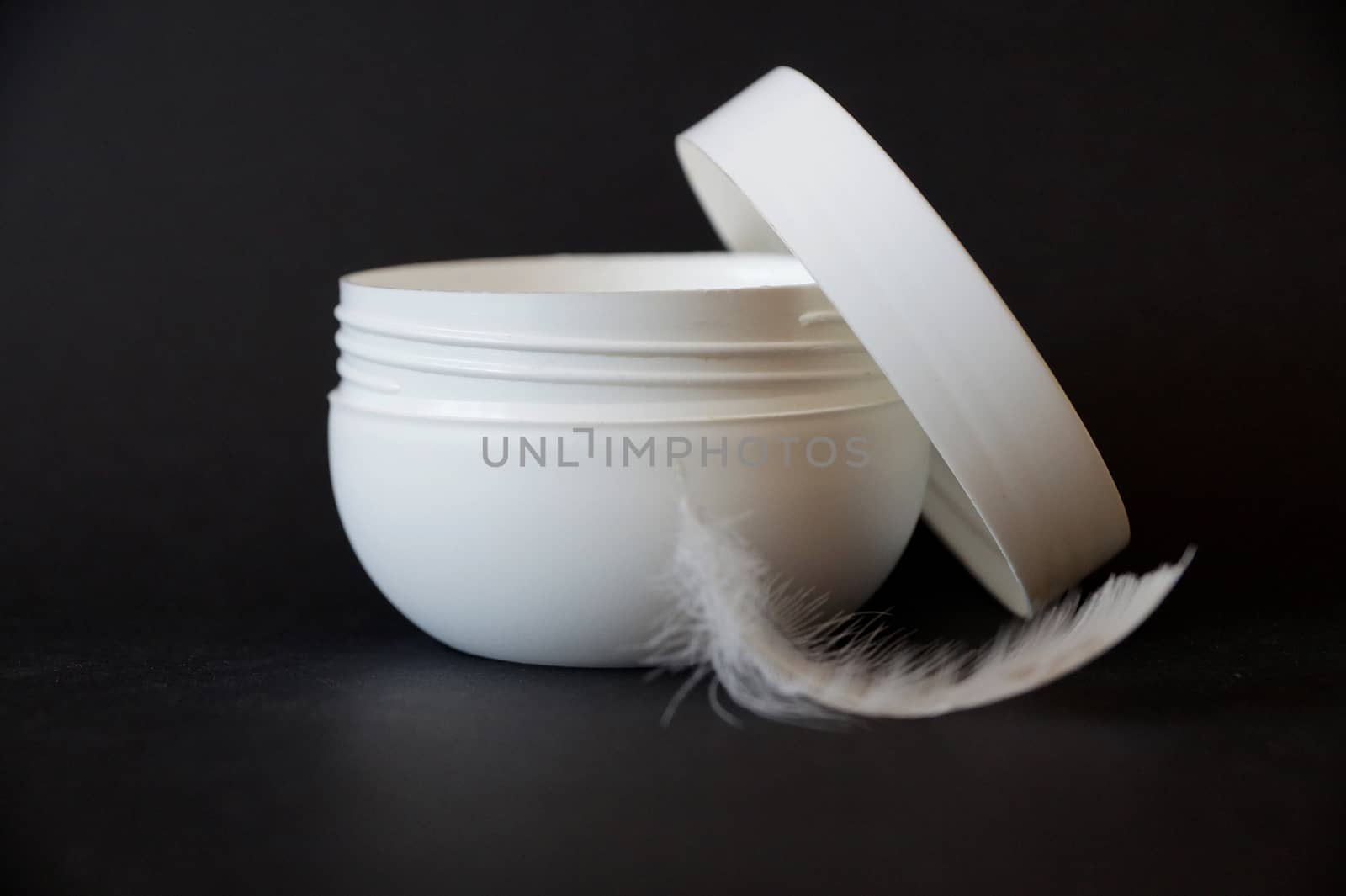 open white jar of cream and white feather on black background, copy space