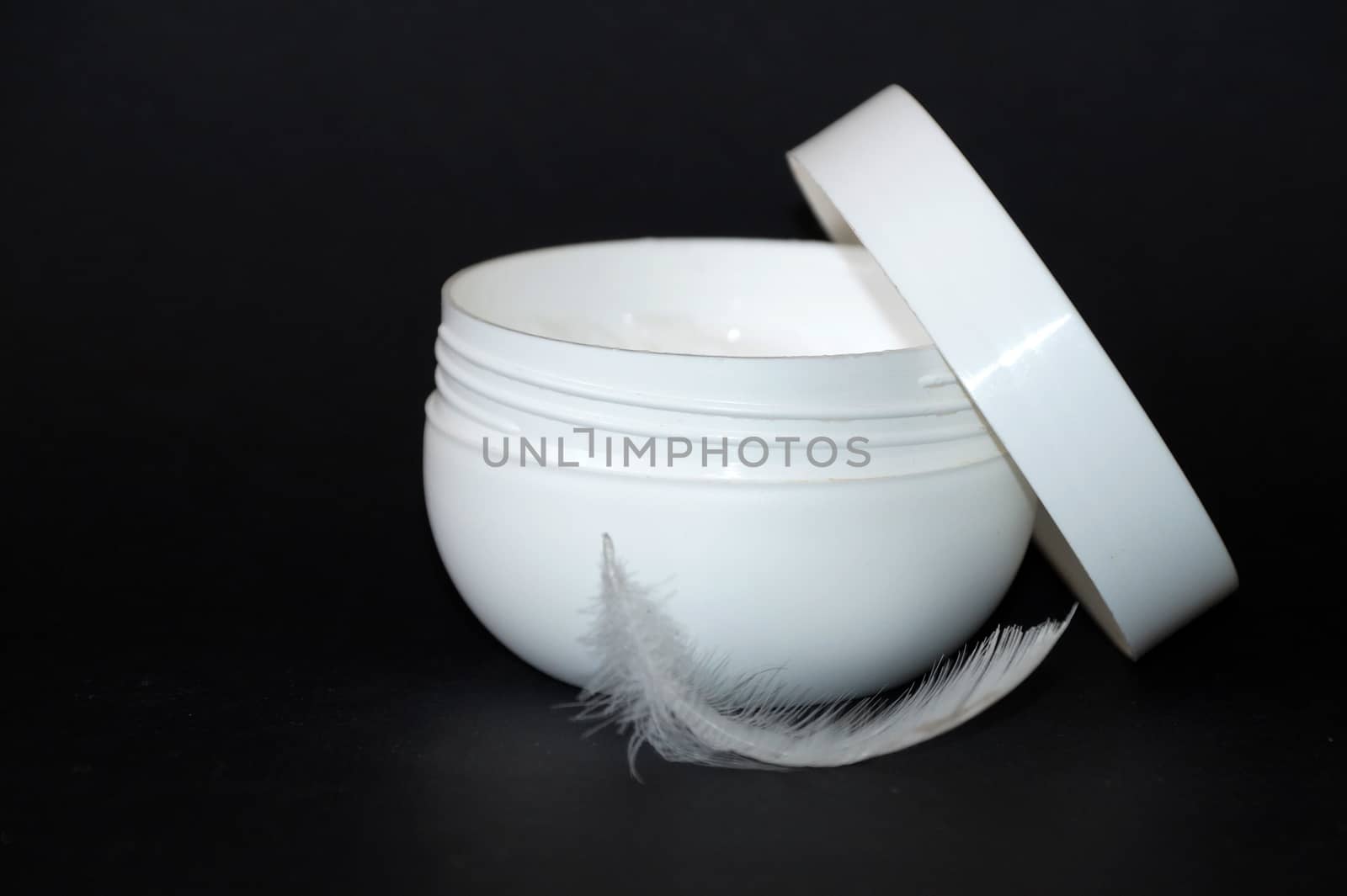 open white jar of cream and white feather on black background by Annado