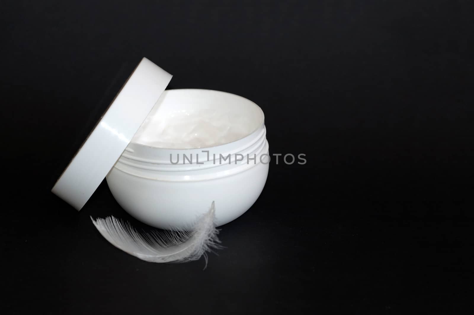 open white jar of cream and white feather on black background by Annado