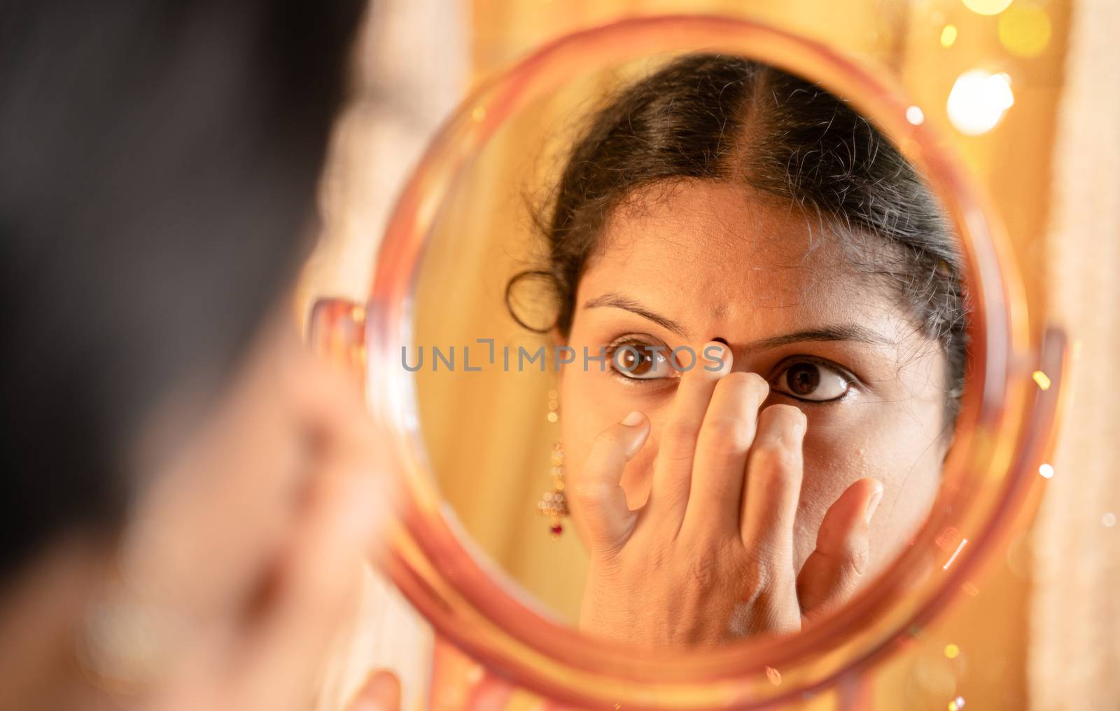 Indian married Woman applying Bindi, sindoor or decorative mark to forehead in front of mirror during festival celebrations with decoration lights as background. by lakshmiprasad.maski@gmai.com