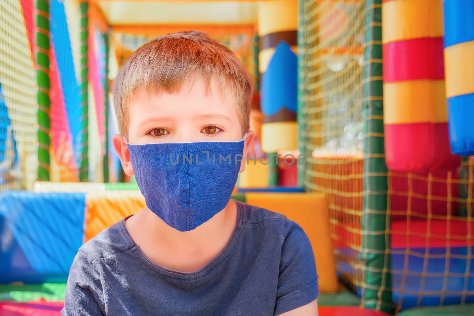 Boy in a medical mask standing near a closed playground. Child in face mask in front of playground . Free play as part of the educational process during covid-19 global pandemic. Coronavirus outbreak by synel