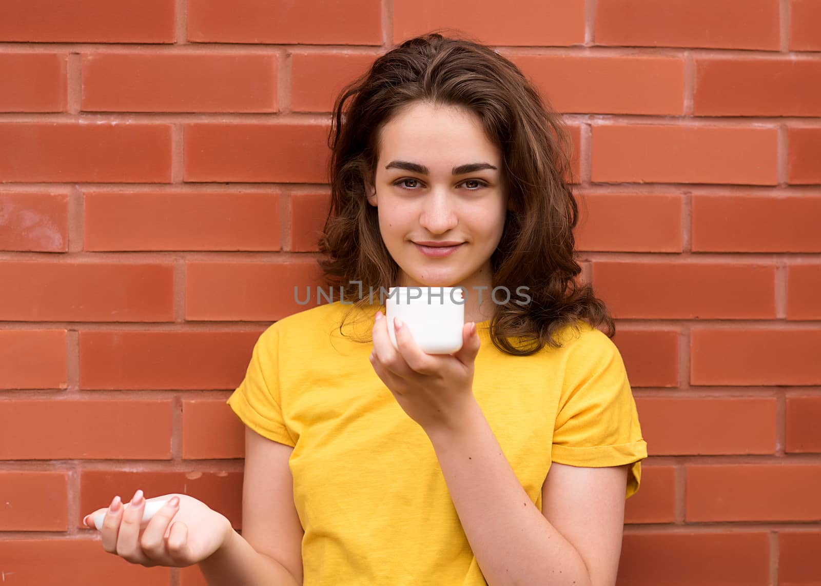 young woman wearing a yellow shirt, looking at a tub of cream against a brick wall