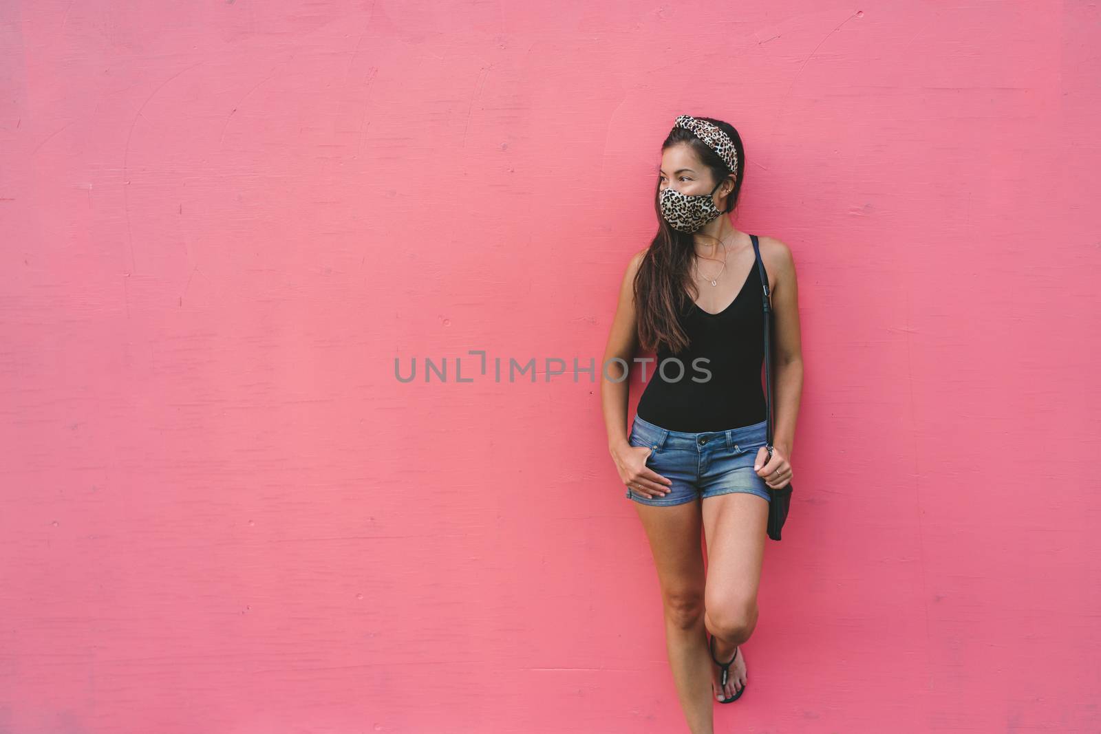 Asian woman leaning on pink wall wearing fabric face mask in fashion leopard pattern print. Casual young people city lifestyle during corona virus by Maridav