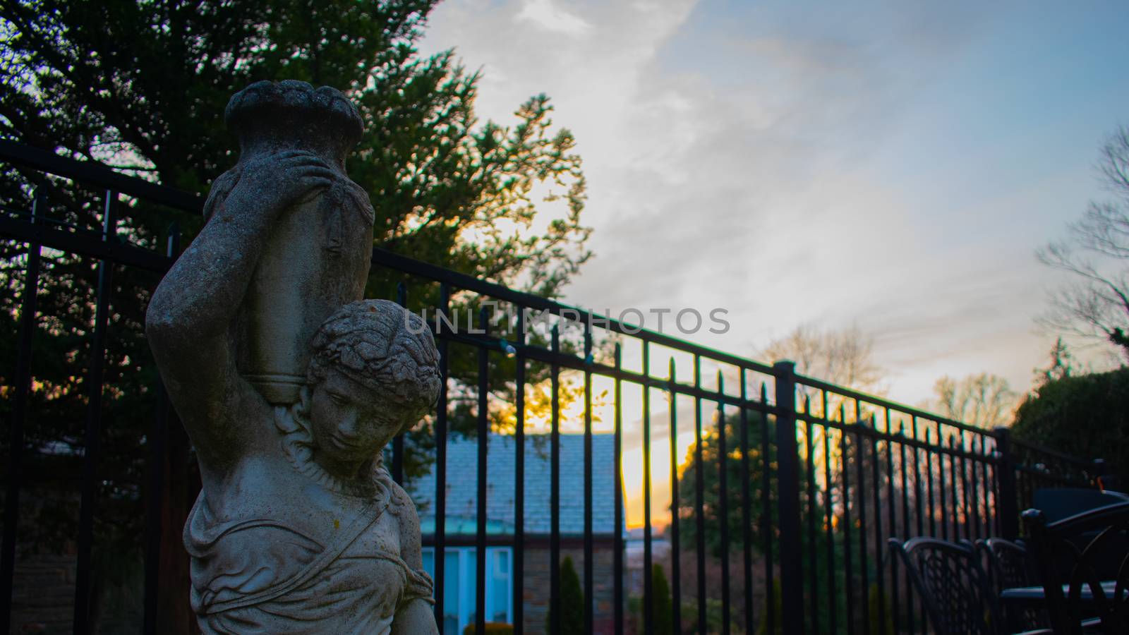 A Statue of a Woman Holding a Vase With a Fence and Sunset in the background