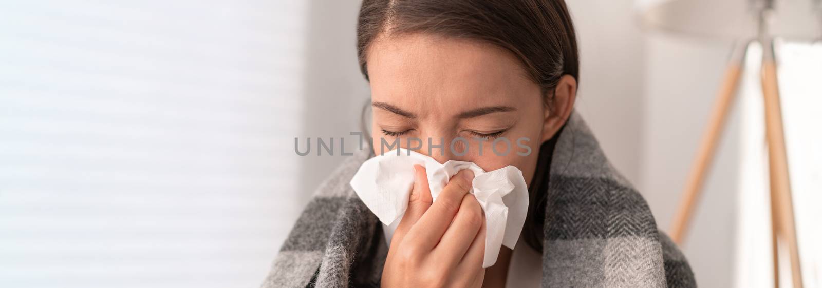 Sick woman feeling unwell staying home. Young girl with flu symptoms coughing in tissue covering nose when sneezing as COVID 19 prevention. Panoramic crop by Maridav