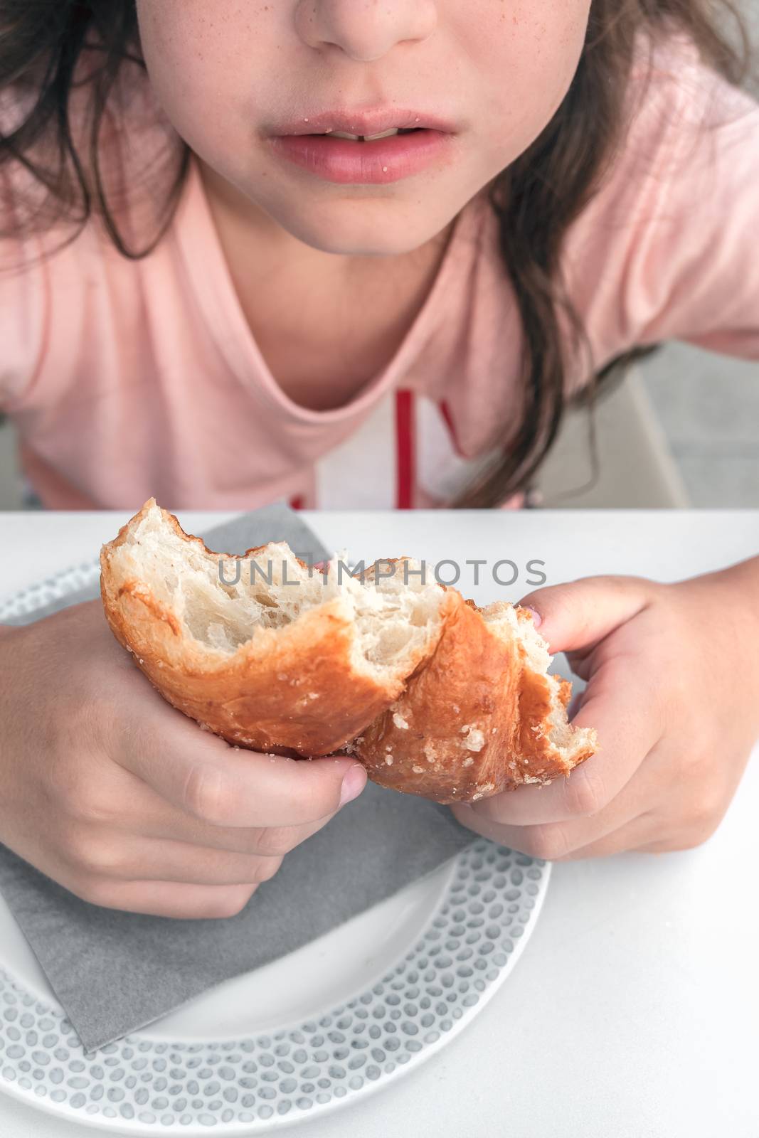 Pretty little girl eats a fresh croissant for breakfast. Portrait of cute girl with yummy face enjoy eating croissant. Healthy food for children concept.