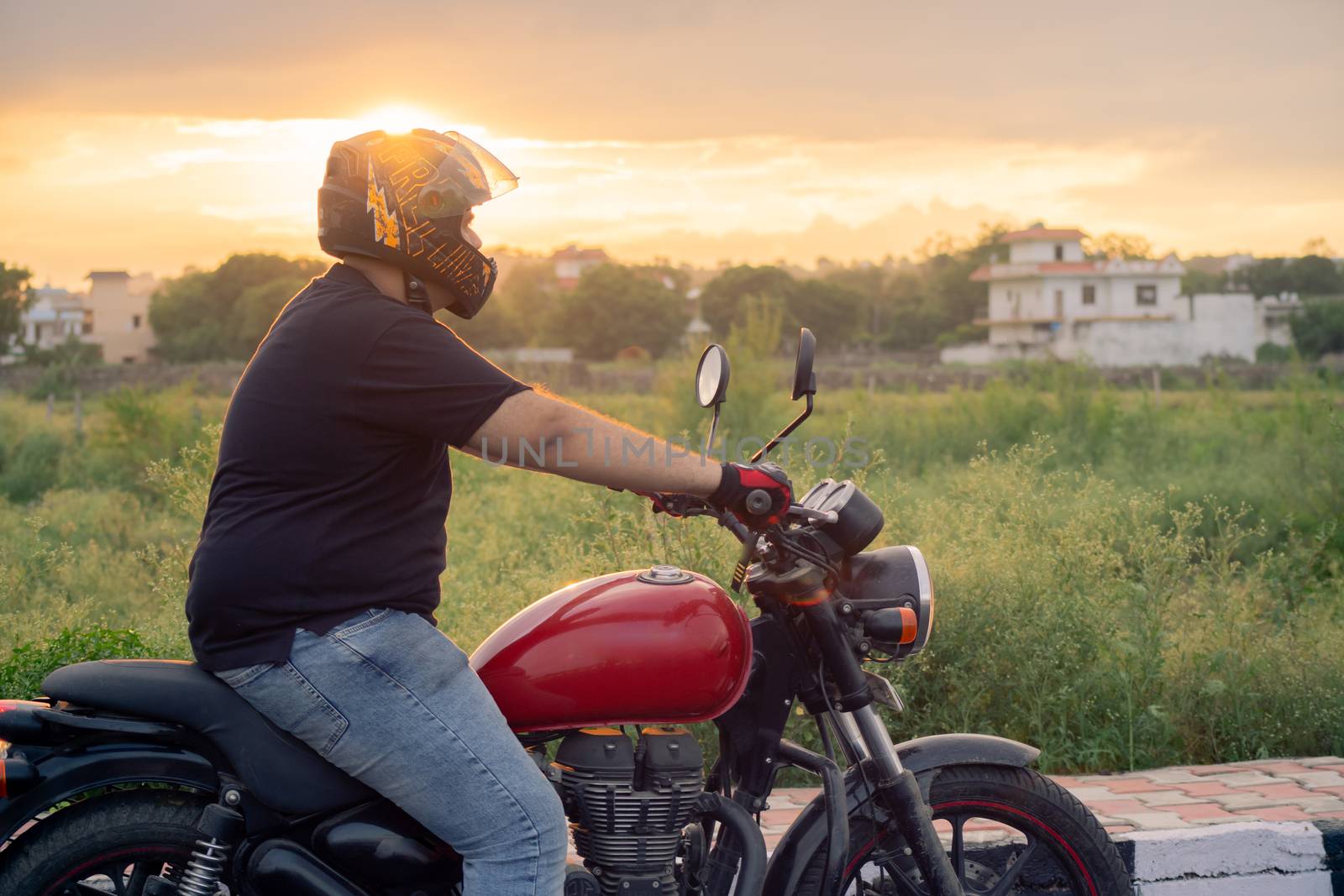 Young indian man with full face helmet and protective gloves riding a red royal enfield at sunset golden hour. Shows the culture of nomadism, travelling and adventure in the beautiful country of india with feilds in the background