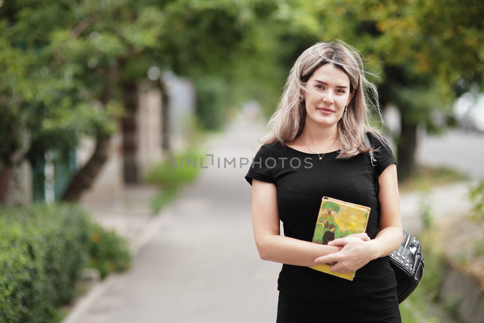 Fashion portrait of young stylish woman walking down the street, in trendy outfit, traveling with a backpack. Holds a book in his hand