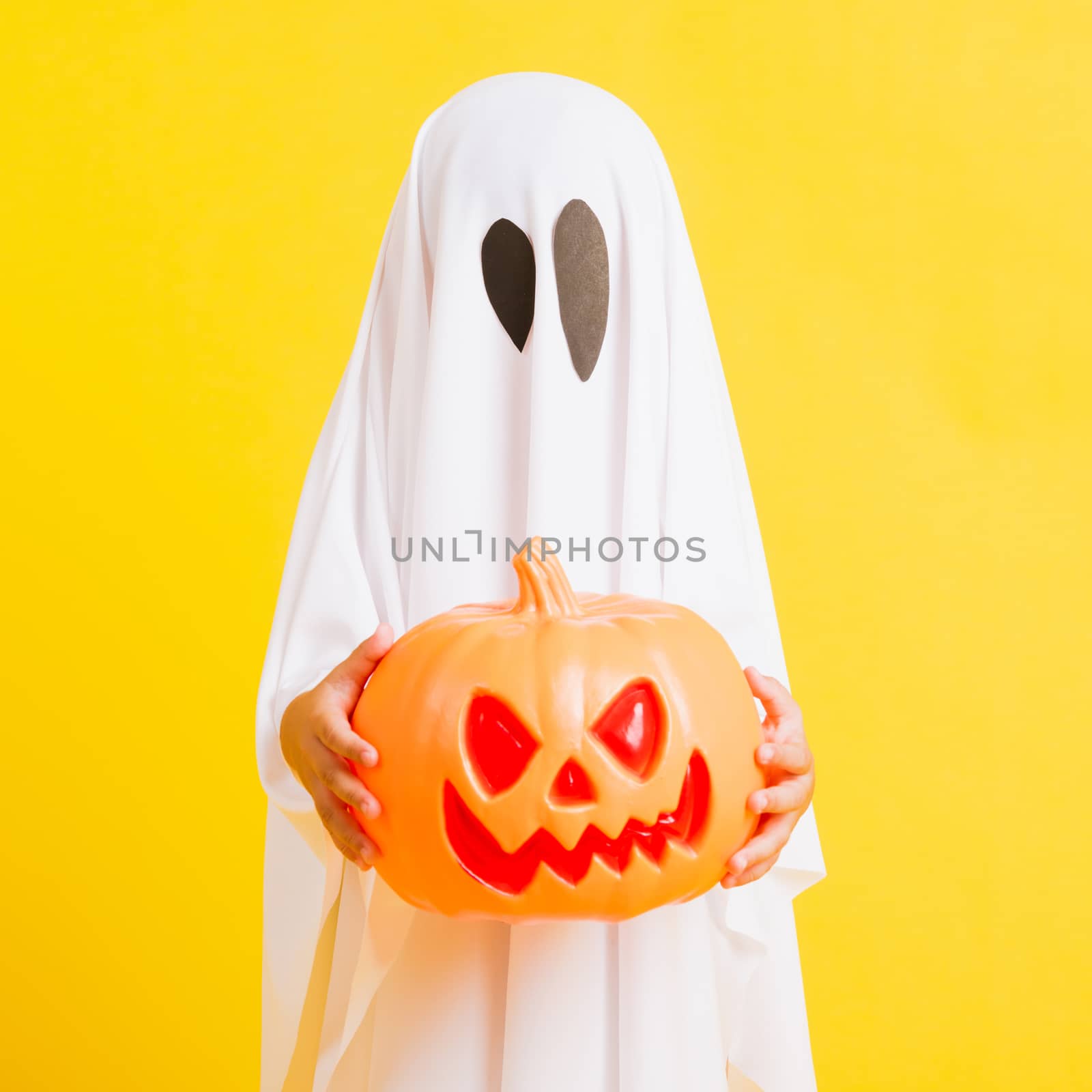 Funny Halloween Kid Concept, little cute child with white dressed costume halloween ghost scary he holding orange pumpkin ghost on hand, studio shot yellow on white background