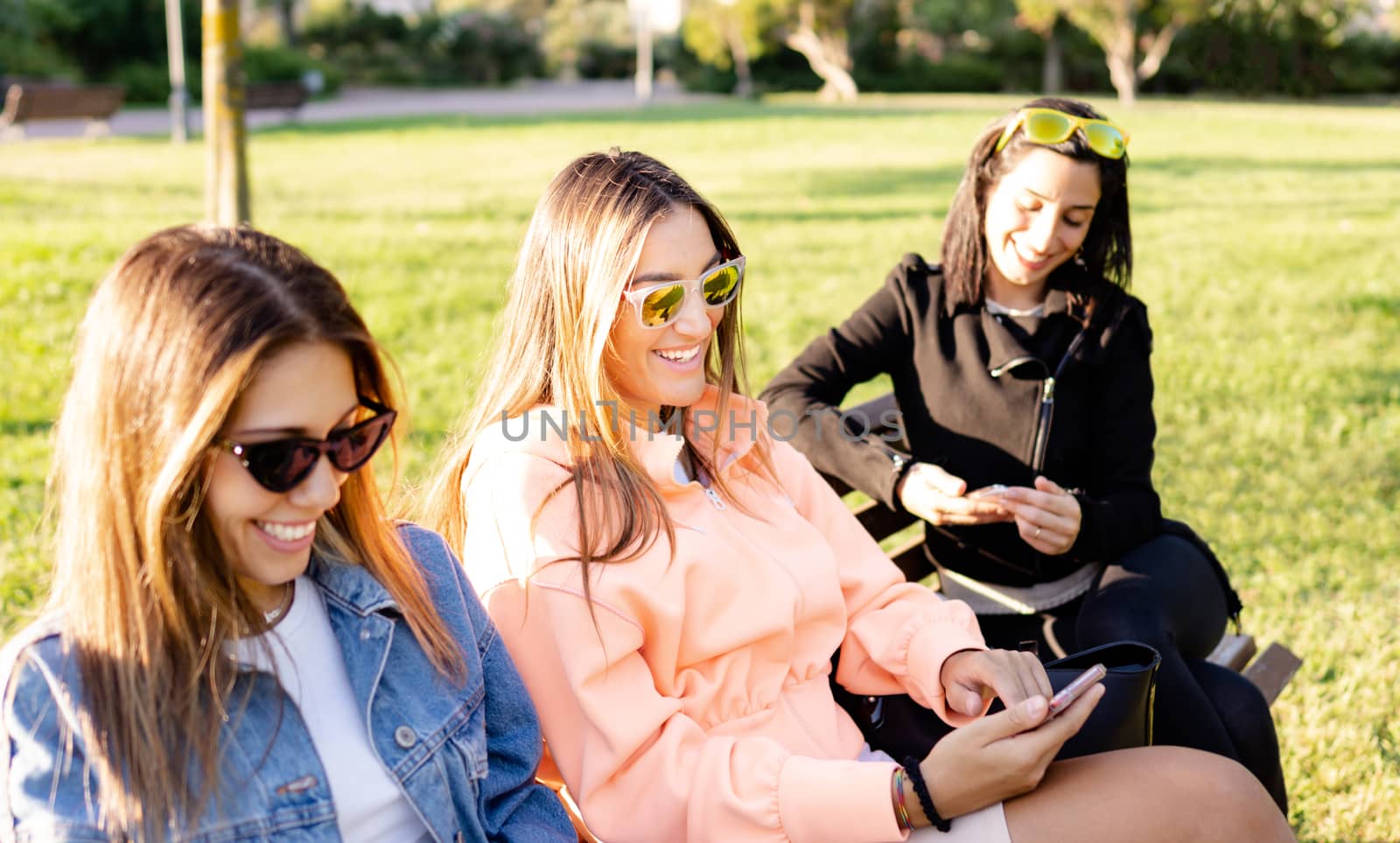Modern young women sitting on a bench in a green parque using smartphone to stay connected with social network - Female friends are shopping online surrounded by nature in contrast with technology by robbyfontanesi